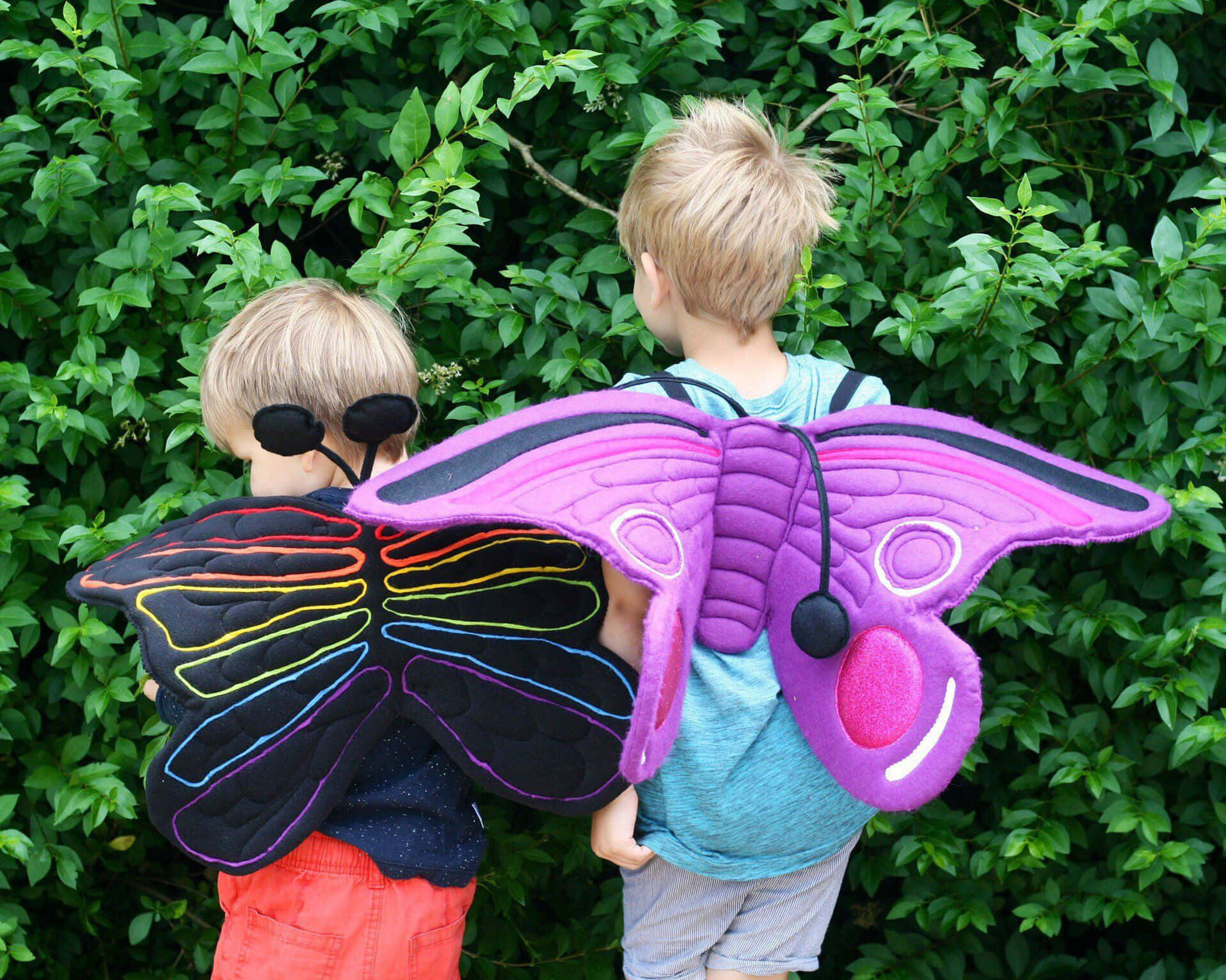Butterfly Wing Dress 桌布 壁紙 — 兩個孩子穿著色彩繽紛的毛絨飛蛾翅膀，背對背站在茂密的綠色籬笆前。