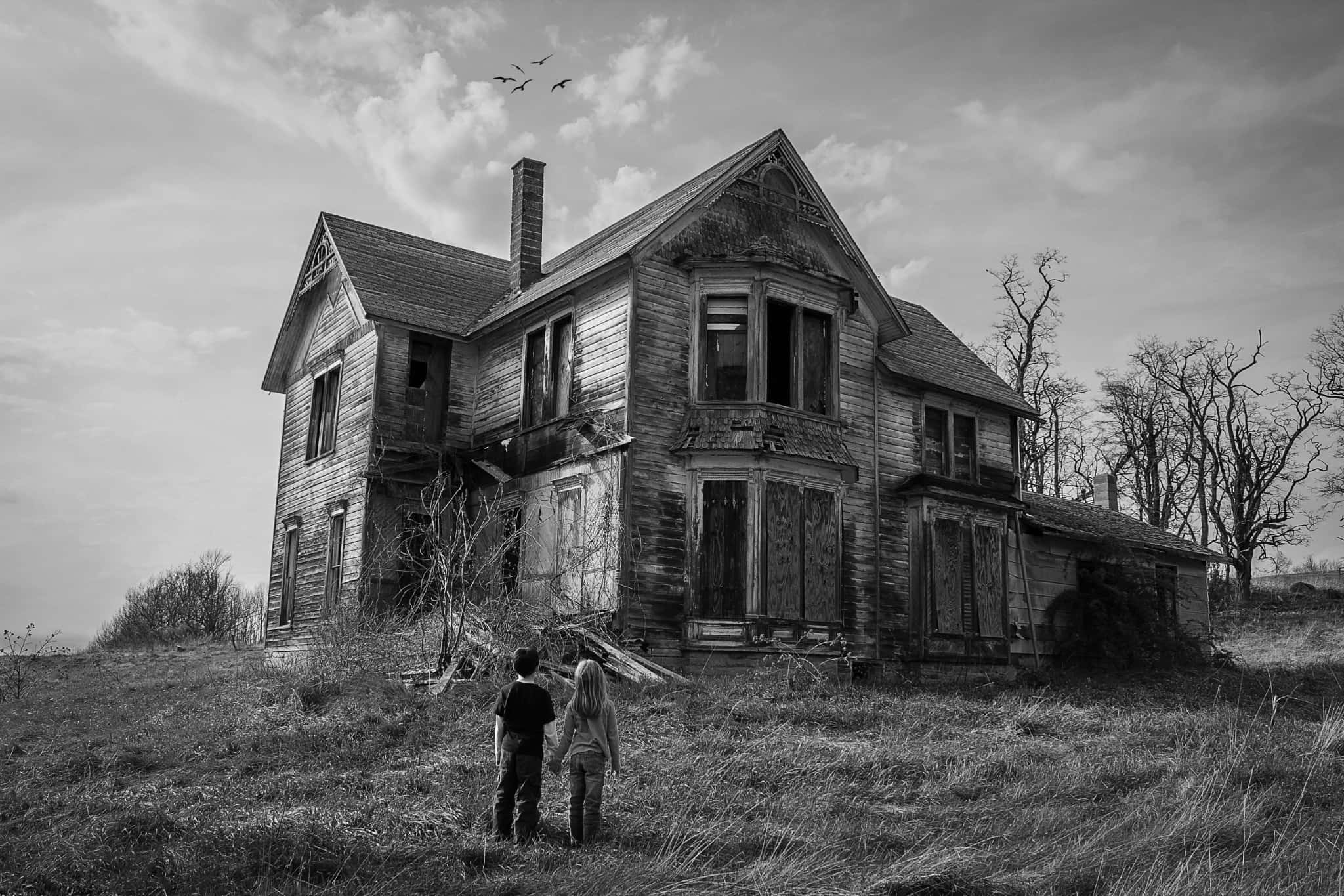 Haunted House 桌布 壁紙 — 兩個孩子站在一座破敗的維多利亞式房屋前，以單色呈現，周圍是光禿禿的樹木和雜草叢生的草地，天空多雲。