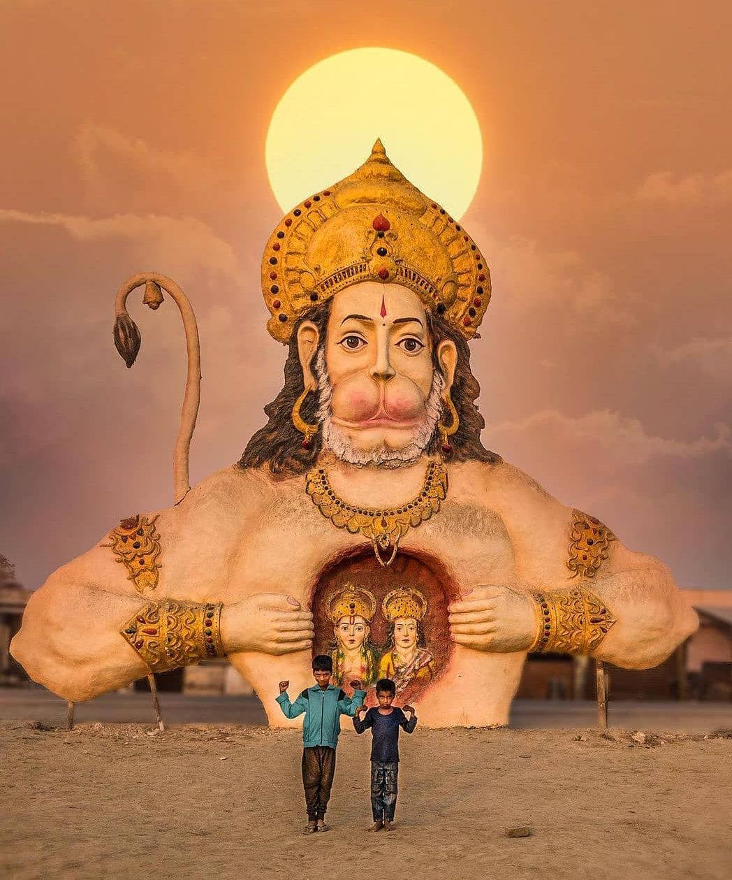Hanuman 桌布 壁紙 — 兩個孩子站在巨大的哈努曼雕像前，身後夕陽西下，雕像胸前懷抱著兩個較小的神祇。