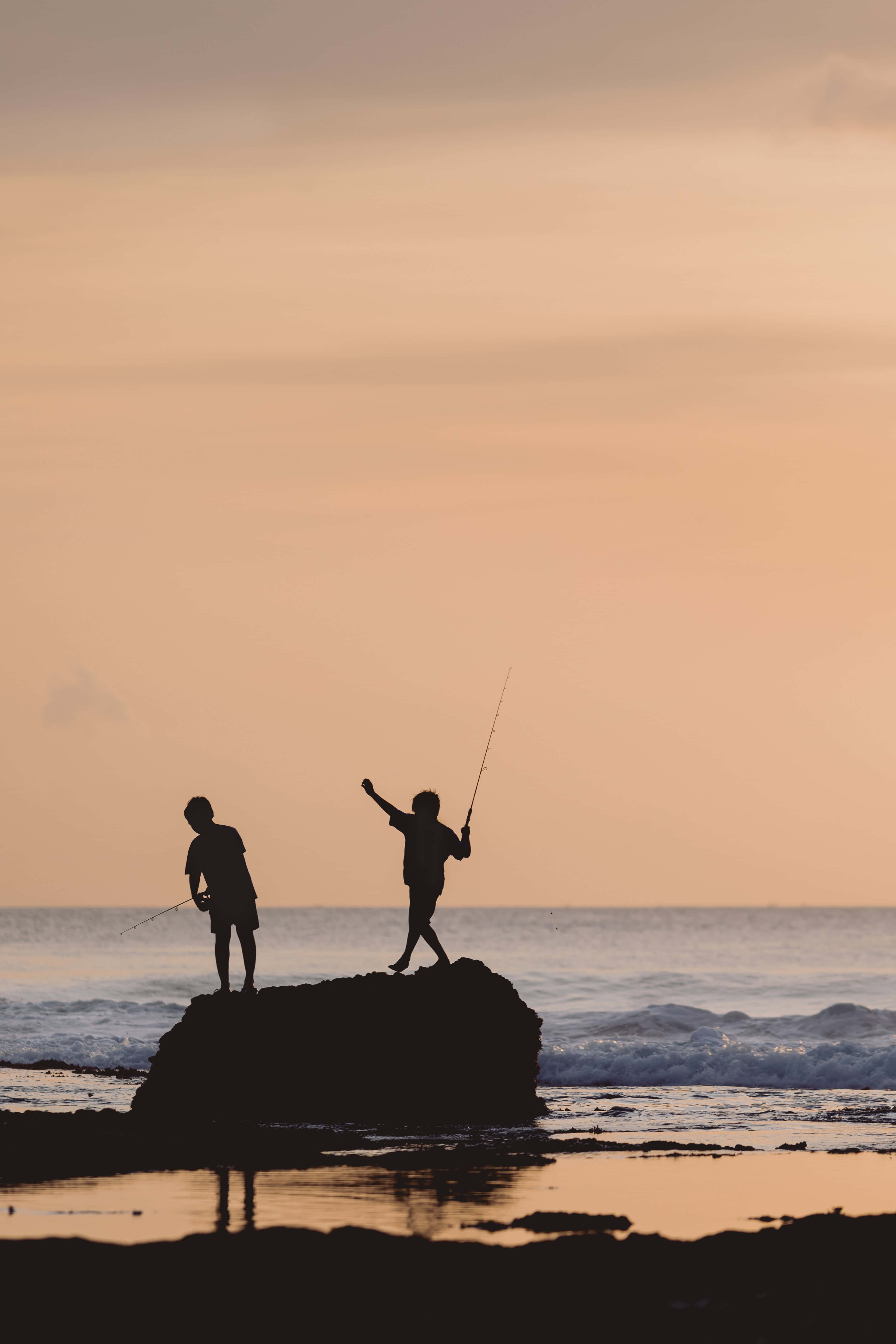 Fishing Phone 桌布 壁紙 — 兩個孩子背對溫暖的夕陽天空，站在海邊岩石上，將釣竿拋向海浪中。