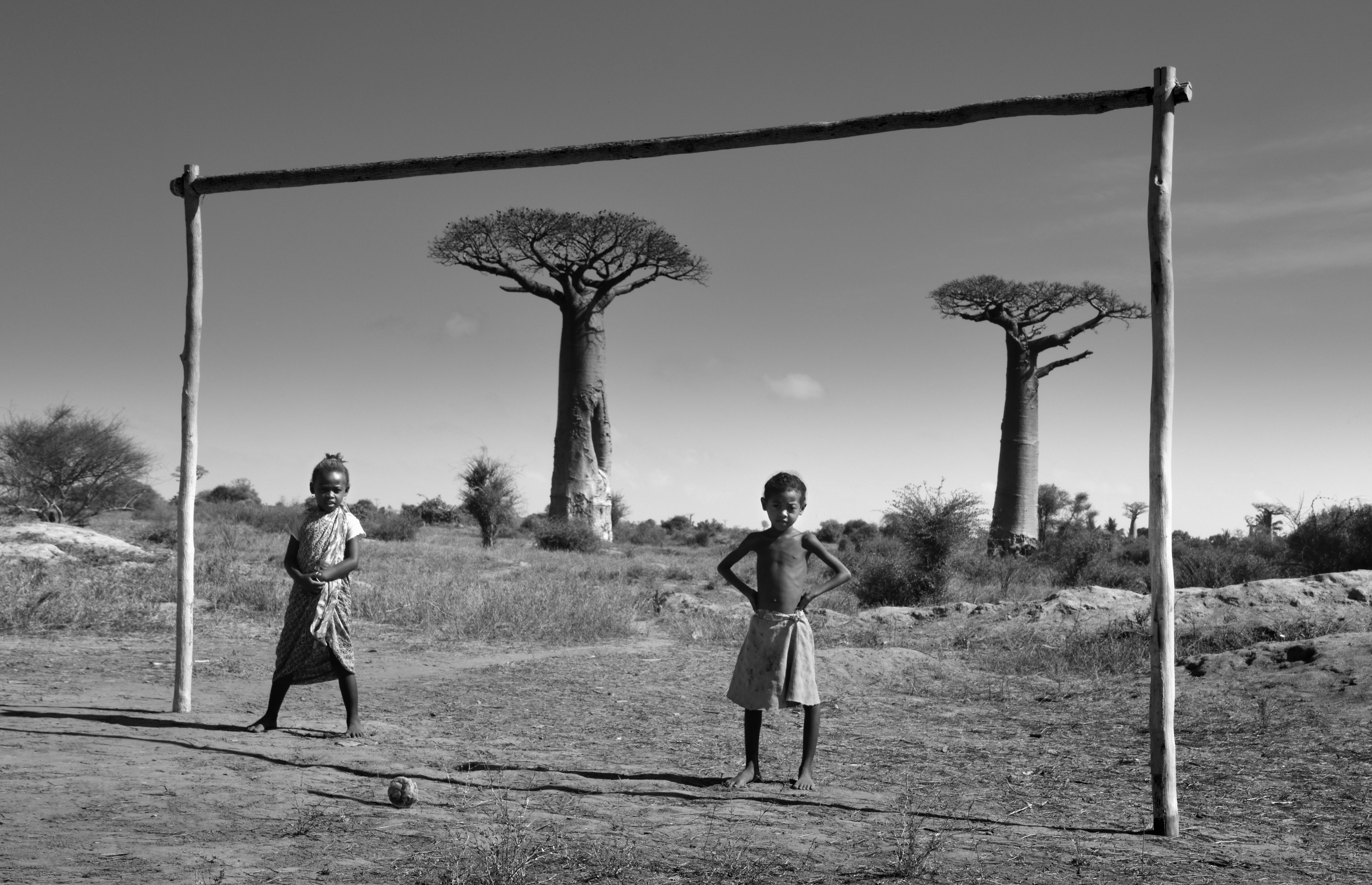 Madagascar 桌布 壁紙 — 兩個孩子自豪地站在一個手工製作的足球球門前，背景是乾燥的稀樹草原，點綴著標誌性的猴麵包樹，頭頂是廣闊的天空。