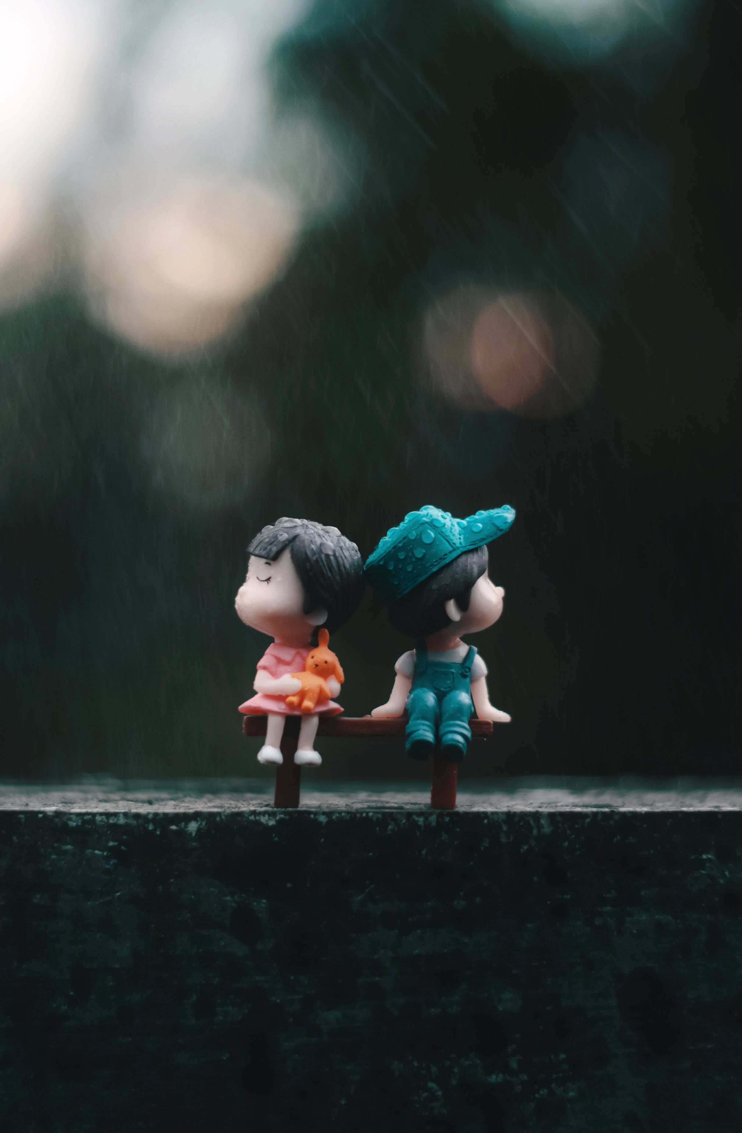 cute doll couple 桌布 壁紙 — 兩個小公仔並排坐在長椅上，細雨輕柔，背景有柔和模糊的燈光。