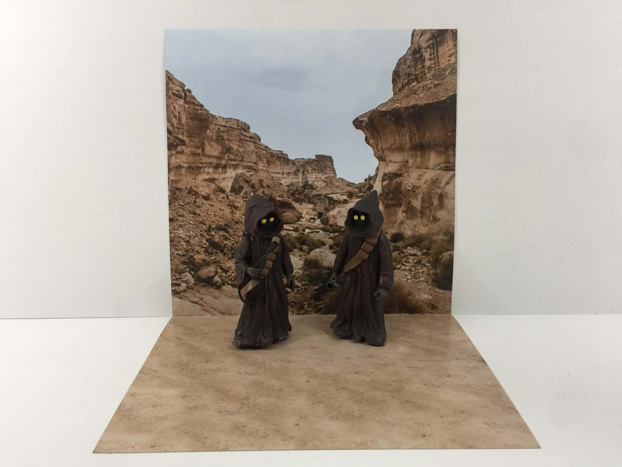 Tatooine 桌布 壁紙 — 兩個小型Jawa人偶站在沙漠峽谷背景前，置於有紋理的紙板底座上。