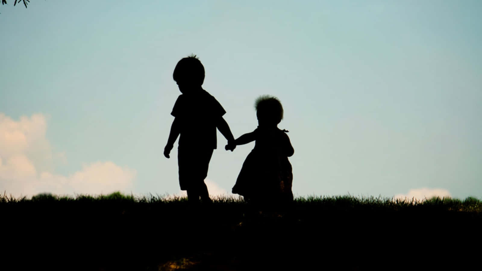 Brother And Sister 桌布 壁紙 — 兩個小孩在柔和的天空下剪影，手牽手走過黃昏時分的草地山丘。