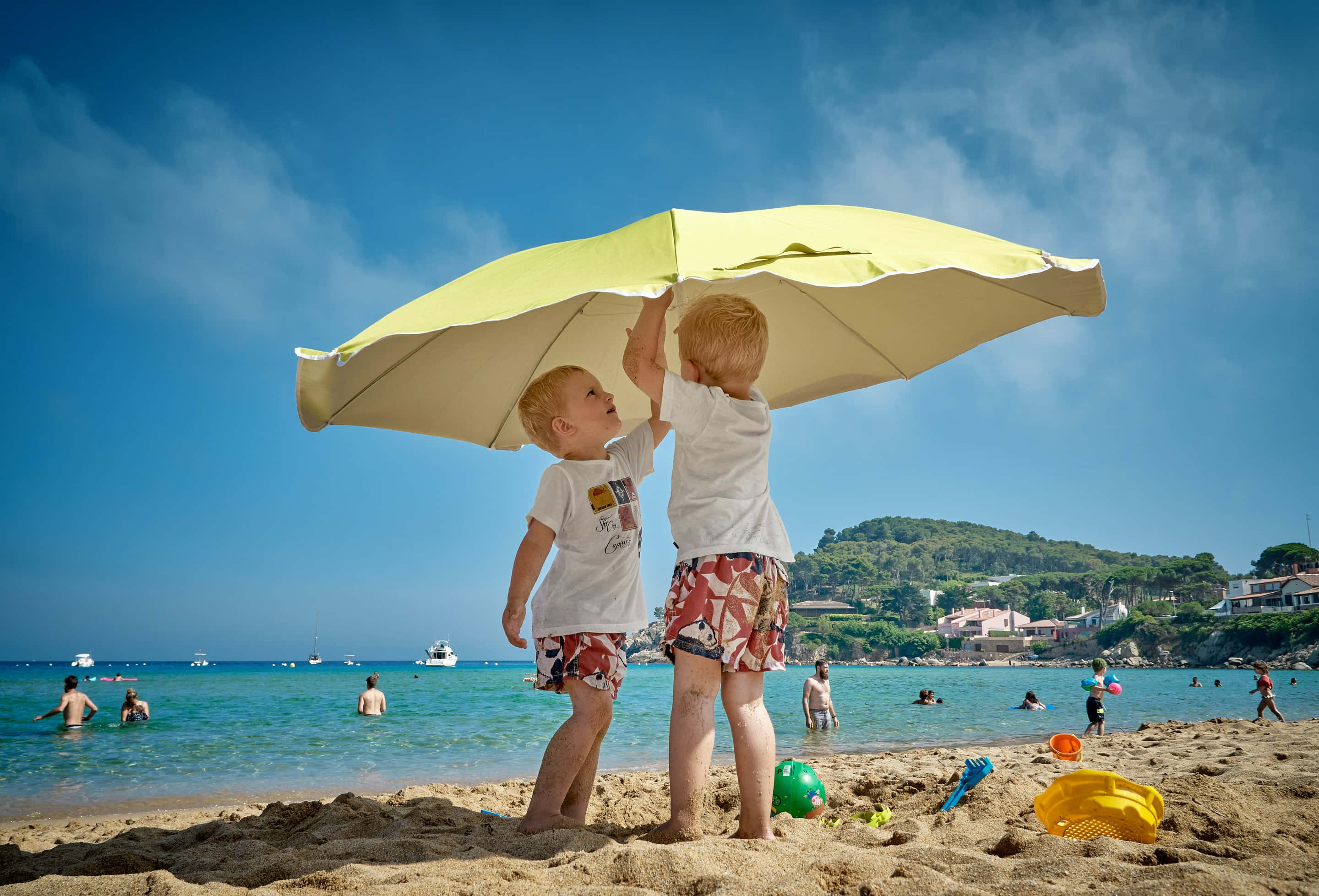 Summer Vacation 桌布 壁紙 — 兩個小男孩站在陽光明媚的海灘上，舉起一把黃色雨傘遮住頭頂，在海邊玩耍。