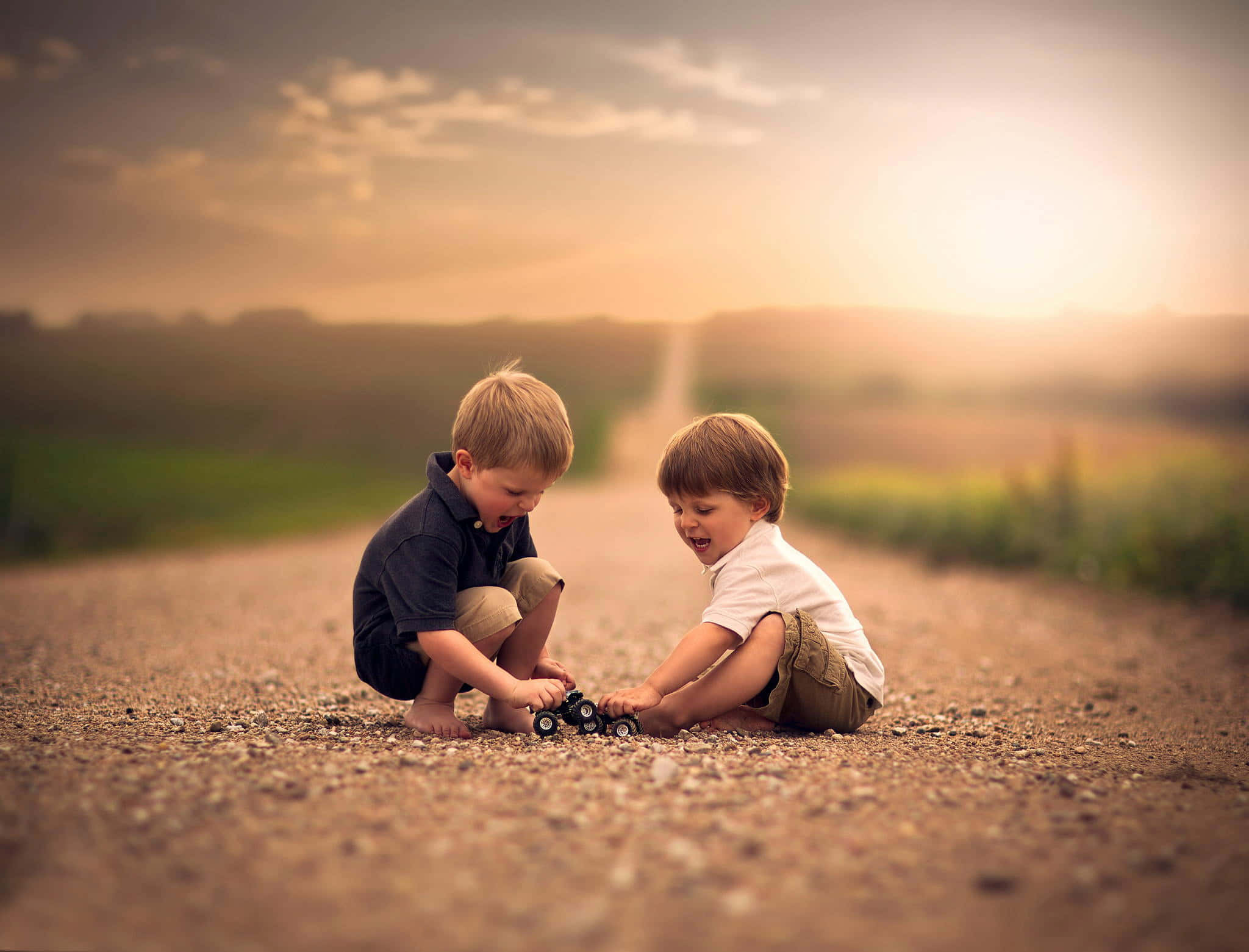 Cute Toddlers 桌布 壁紙 — 兩個小男孩蹲在碎石路上，在金色夕陽下玩玩具車，笑得很開心。