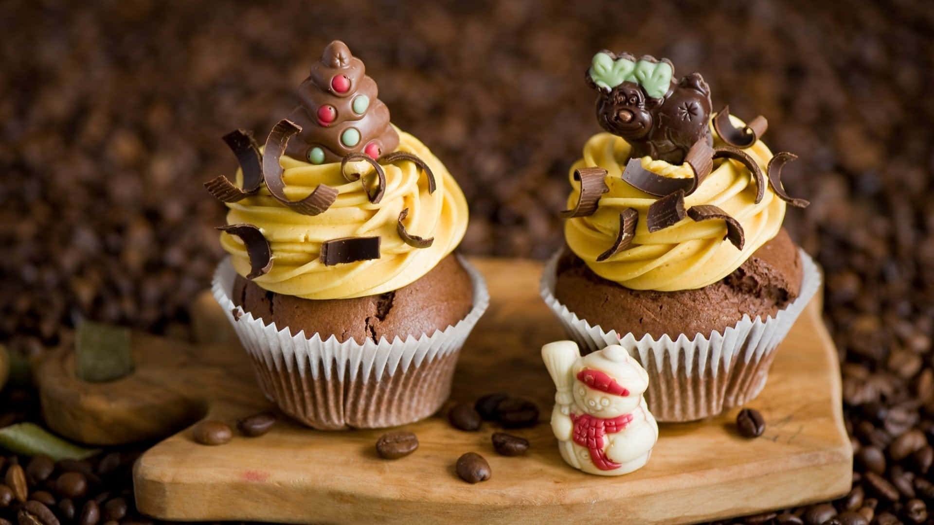 Cupcake 桌布 壁紙 — 兩個巧克力杯子蛋糕，上面有黃色糖霜和巧克力裝飾，放在一個被咖啡豆包圍的木板上。