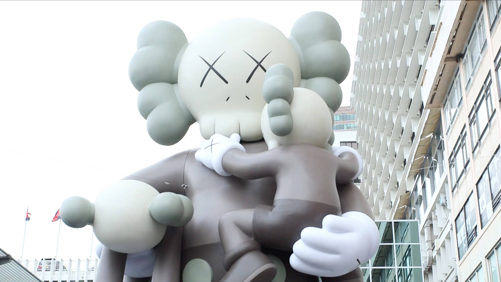 KAWS 桌布 壁紙 — 兩個巨型KAWS公仔在陰天背景下，在現代城市建築物前溫柔擁抱。