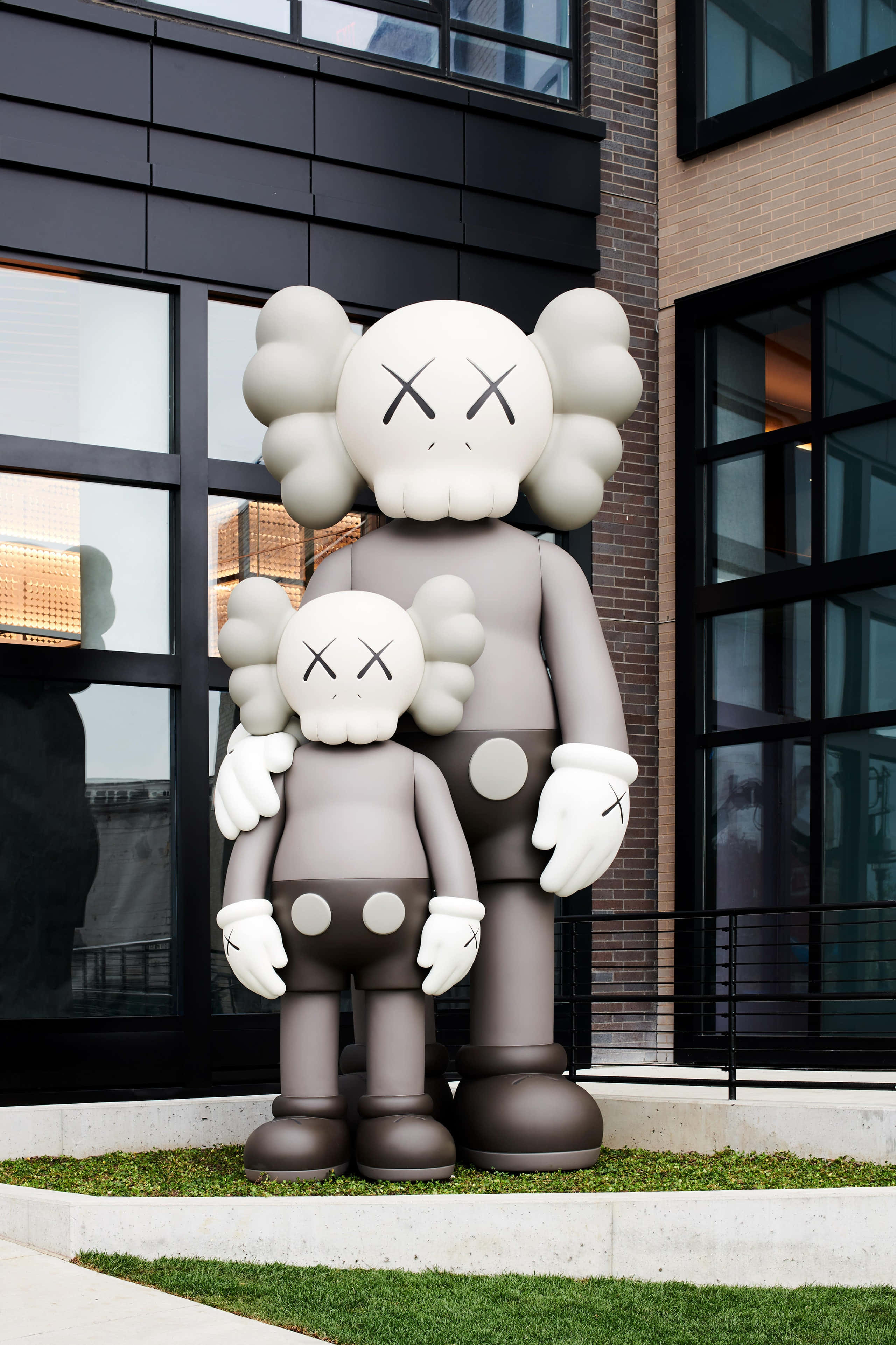 KAWS 桌布 壁紙 — 兩個巨大的灰色KAWS Companion雕像並排站在現代磚和玻璃建築外牆前的草地上。