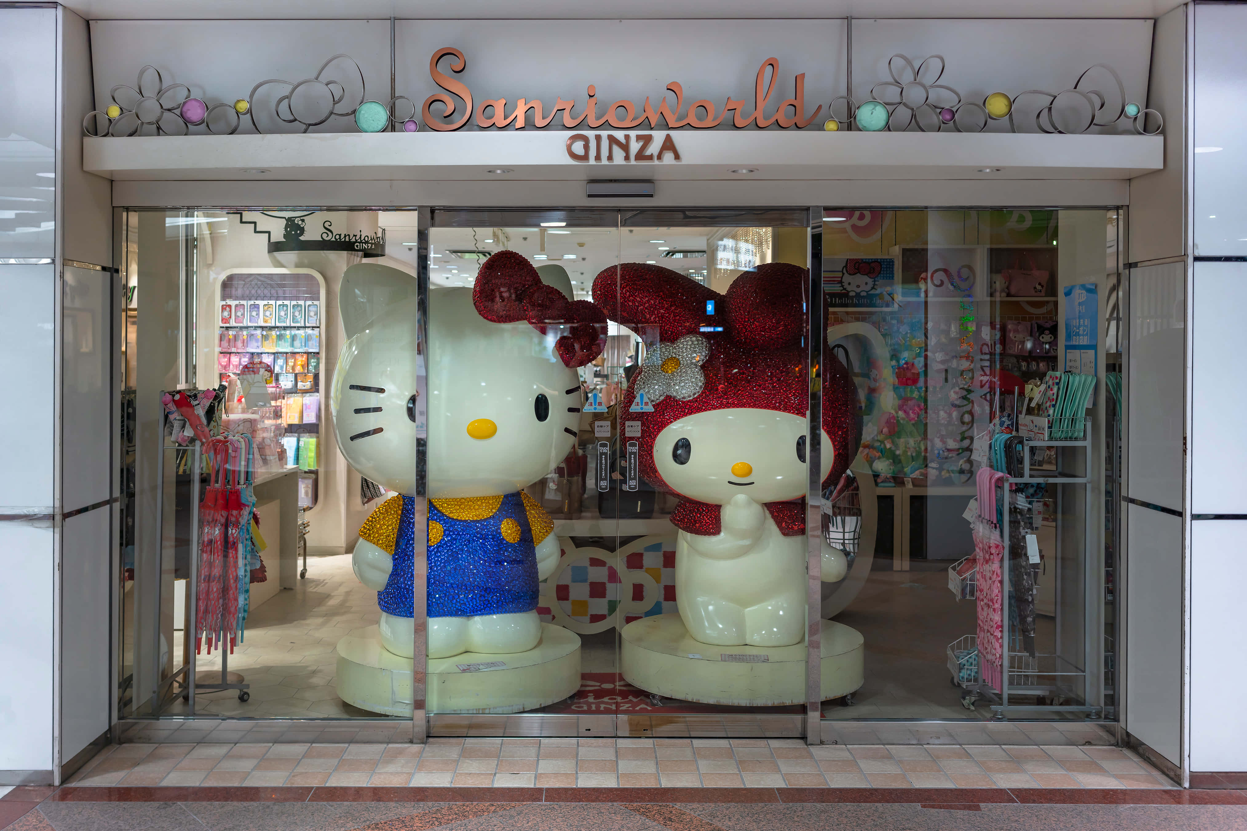 Hello Kitty 桌布 壁紙 — 兩個巨大的閃亮Hello Kitty雕像矗立在Sanrio World Ginza的玻璃入口處，周圍環繞著色彩繽紛的商品。