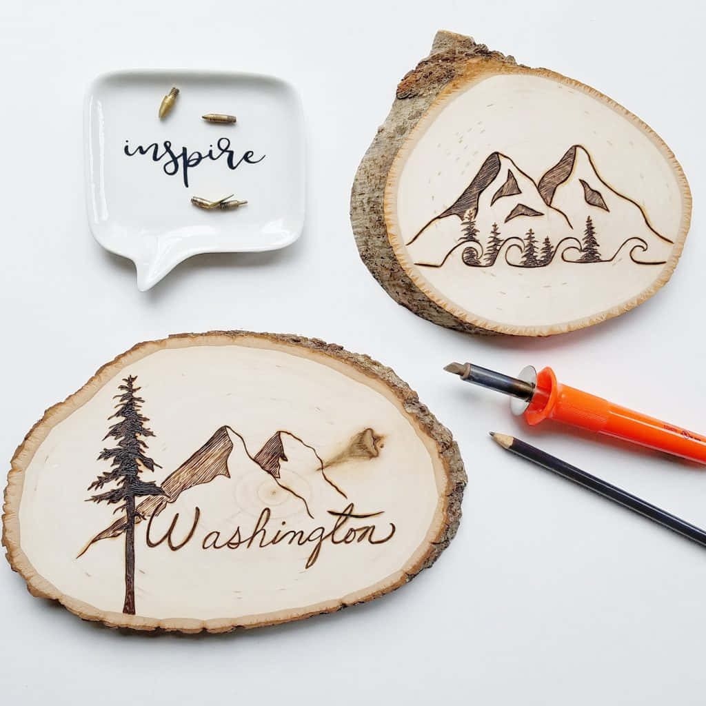 Wood Burning 桌布 壁紙 — 兩個帶有山脈和樹木烙畫設計的木片杯墊，旁邊放著一個帶有 'inspire' 字樣的對話氣泡碟子和工具，置於潔白表面上。