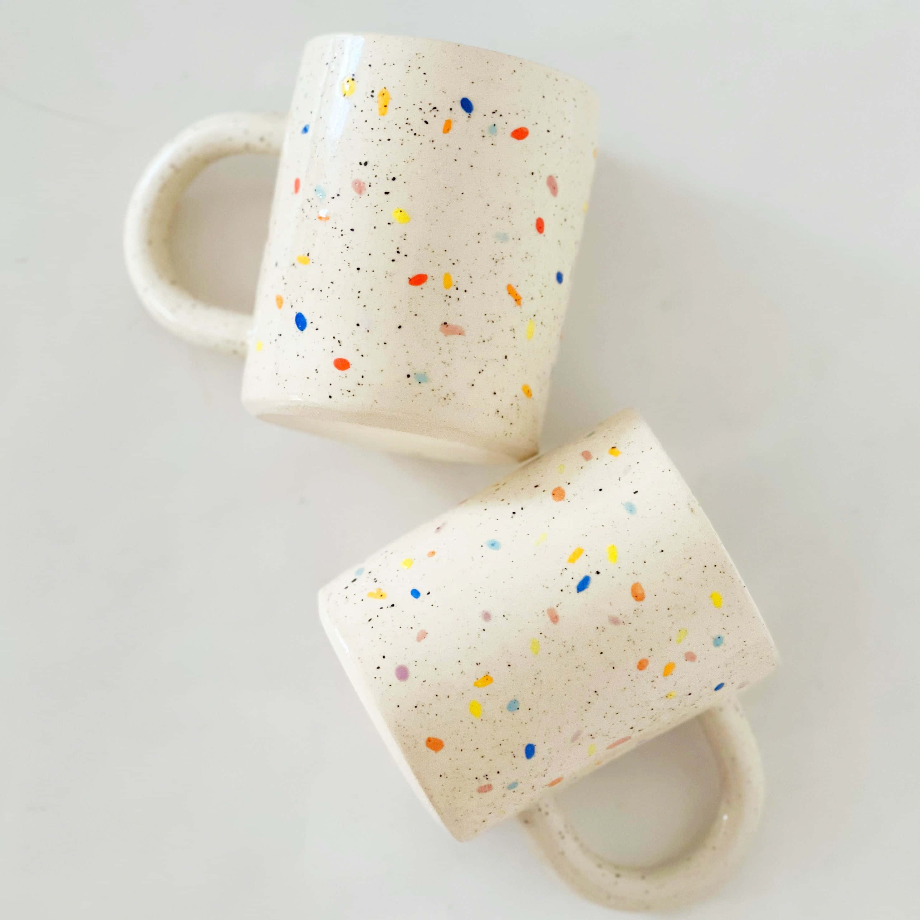 Ceramic 桌布 壁紙 — 兩個帶有彩色圓點的斑點馬克杯放在乾淨的白色表面上，傾斜以顯示其手柄和紋理釉面。