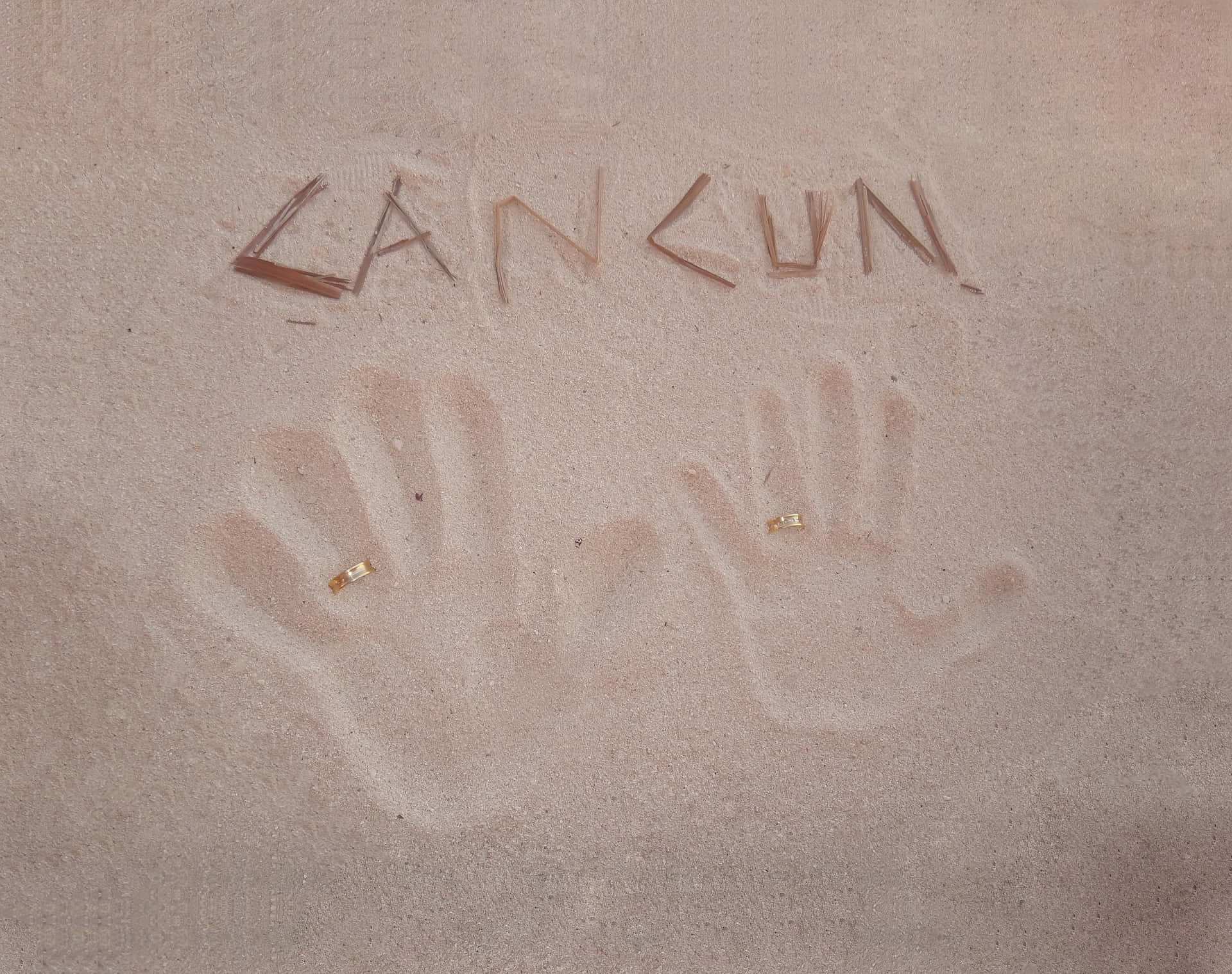 Cancun 桌布 壁紙 — 兩個帶有結婚戒指的手印壓在軟沙上，用乾草字母拼出名字「LANCUN」。