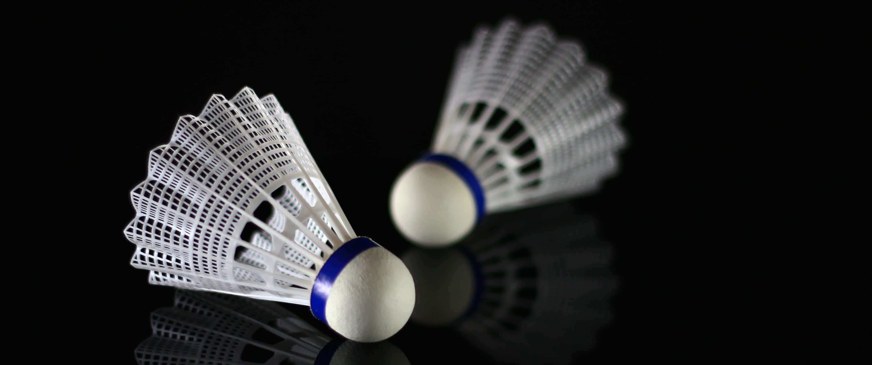 3440x1440p Badminton 桌布 壁紙 — 兩個帶有藍色帶的白色羽毛球在光滑的黑色表面上休息，投射出清晰的倒影。