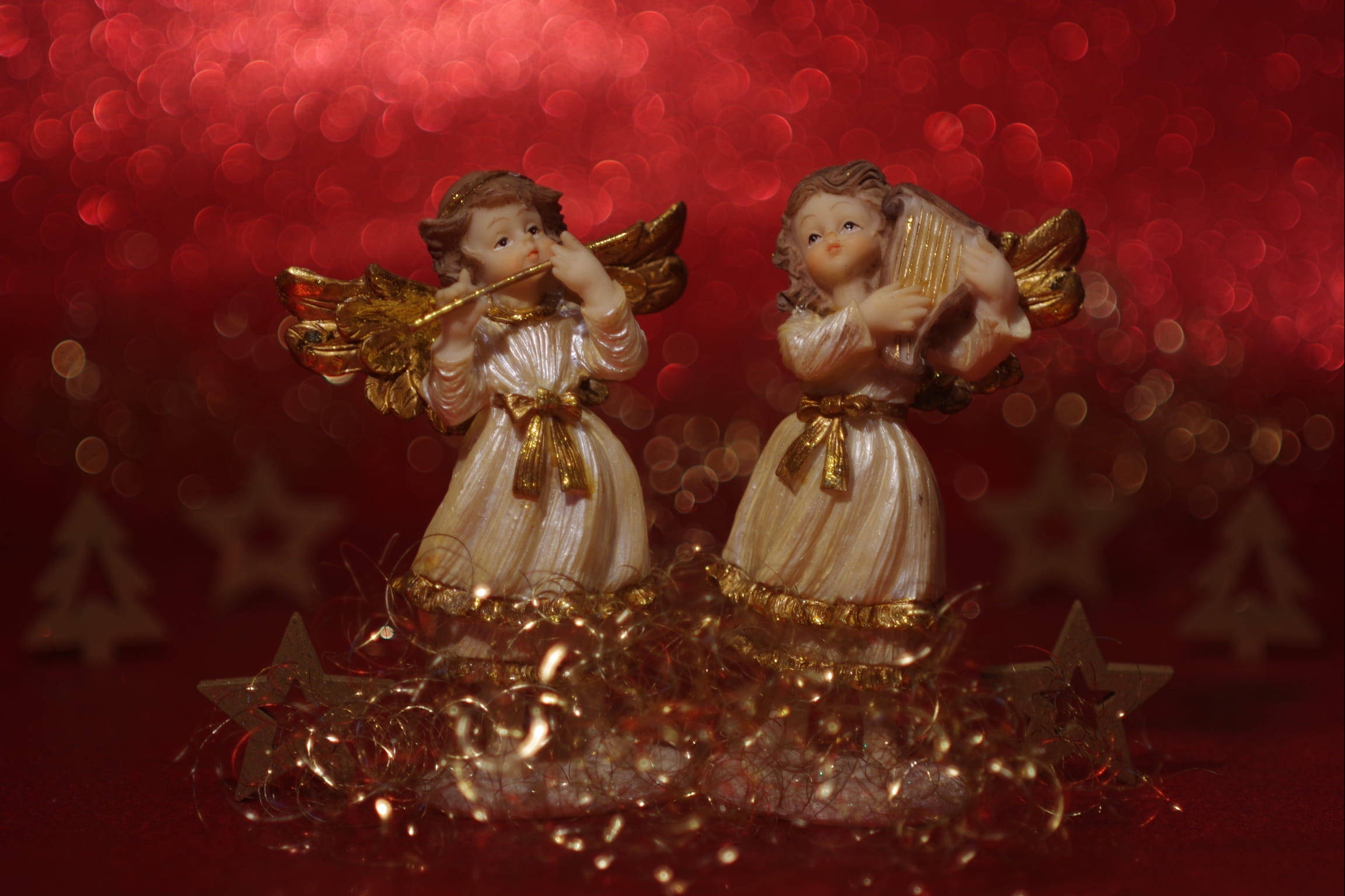 angels 桌布 壁紙 — 兩個帶有金色光環的天使雕像在閃爍的紅色散景背景下演奏樂器，背景有節日星星和閃粉。