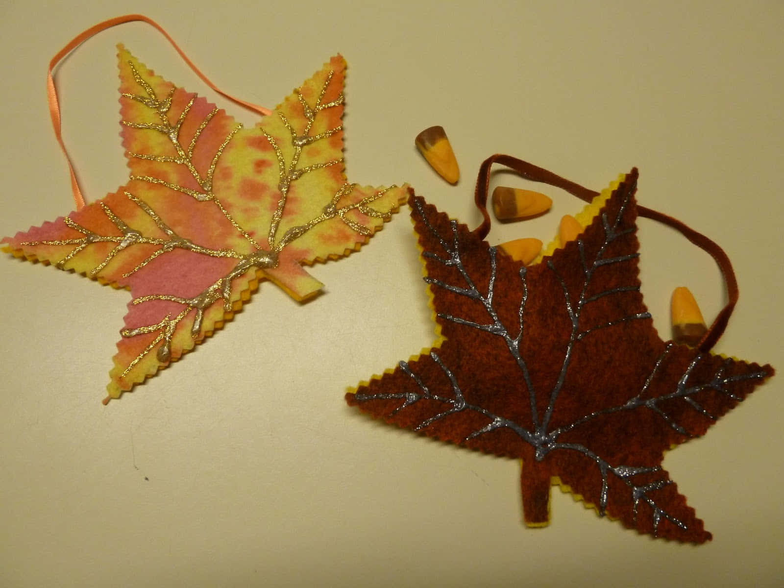 Fall Crafts 桌布 壁紙 — 兩個帶有閃粉葉脈和絲帶環的氈製楓葉裝飾品，放在細小玉米旁邊的純米色表面上。
