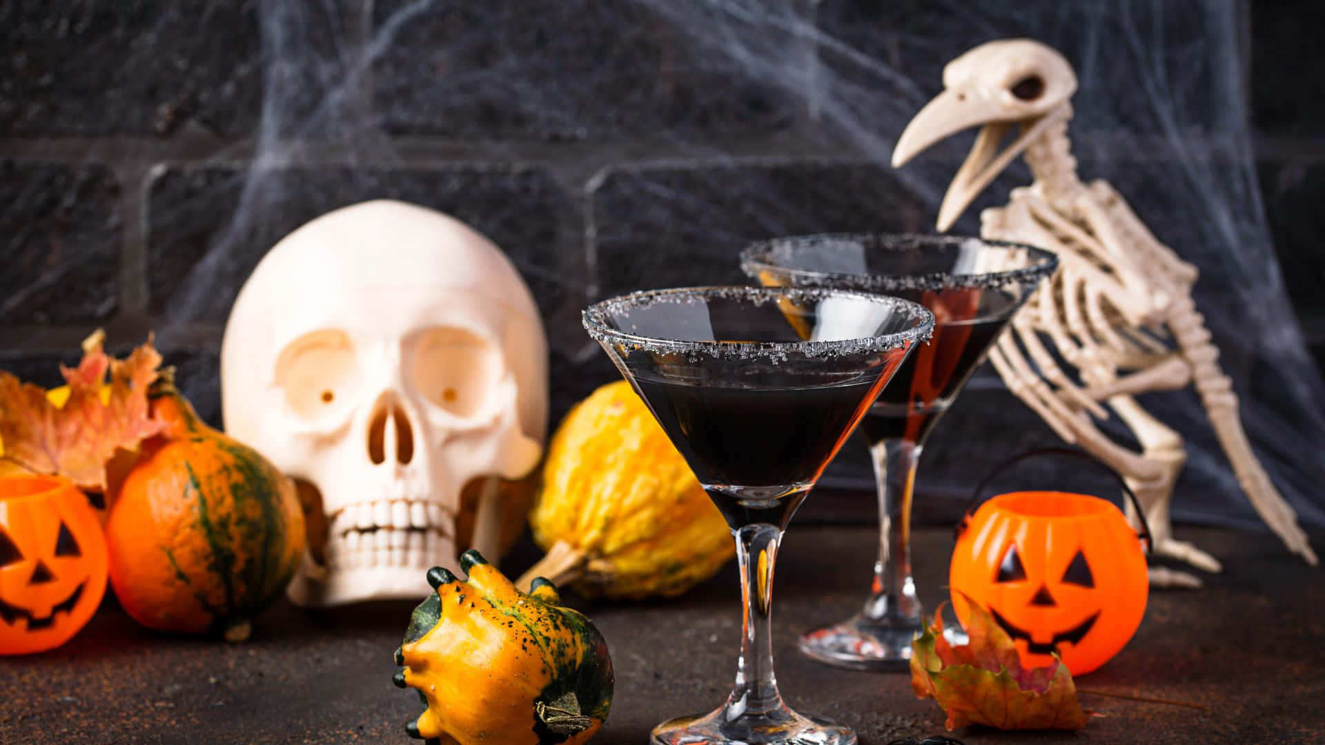 Halloween Cocktails 桌布 壁紙 — 兩個帶有鹽邊的黑色雞尾酒杯放在一個骷髏南瓜和一個骷髏旁邊，背景是深色磚牆。