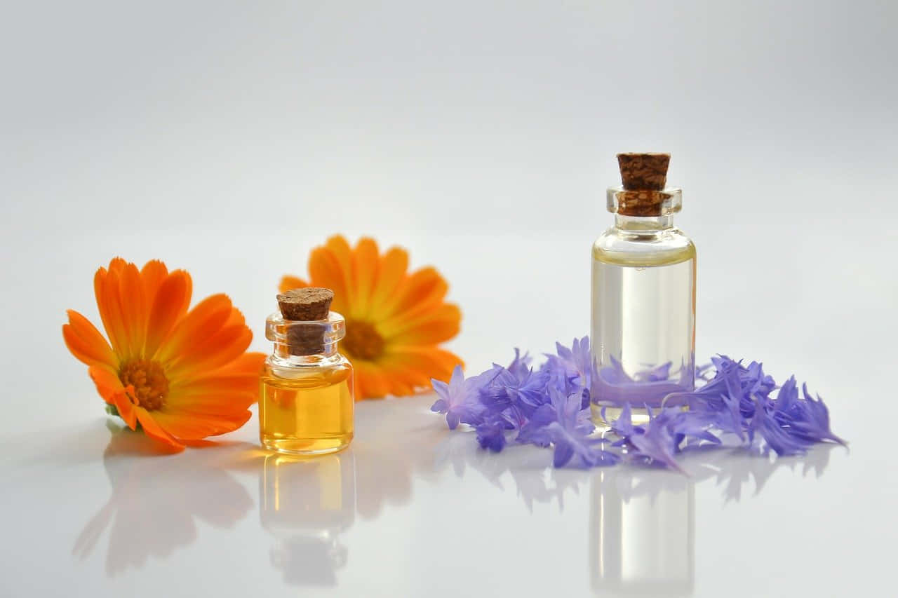 Aromatherapy 桌布 壁紙 — 兩個帶軟木塞的玻璃瓶盛裝著金色和清澈的液體，旁邊是鮮豔的橙色萬壽菊和精緻的紫色花朵，放在反光的白色表面上。