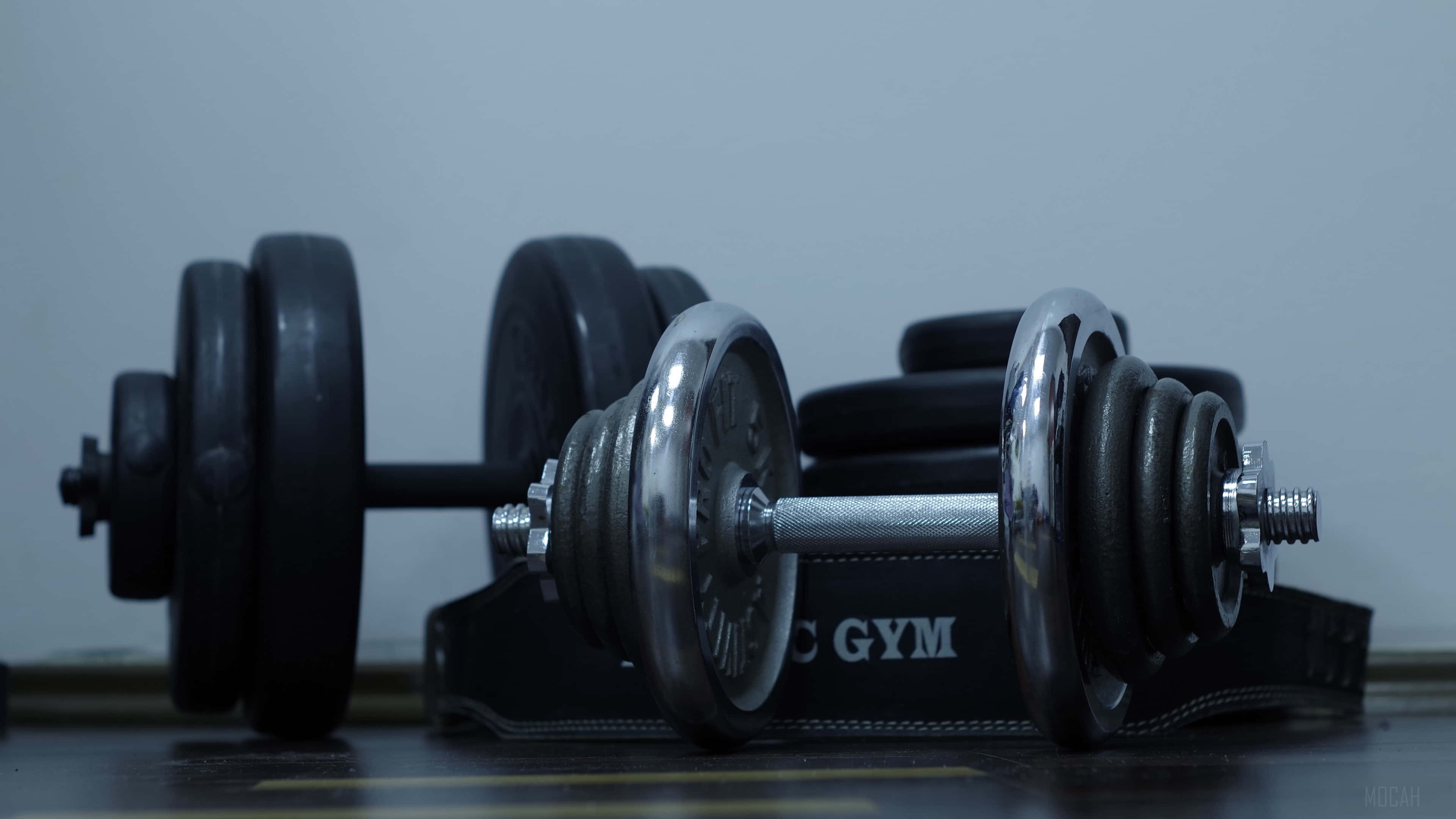 Fitness 桌布 壁紙 — 兩個帶黑色槓鈴片的鉻合金啞鈴放在健身墊上，靠著素牆。
