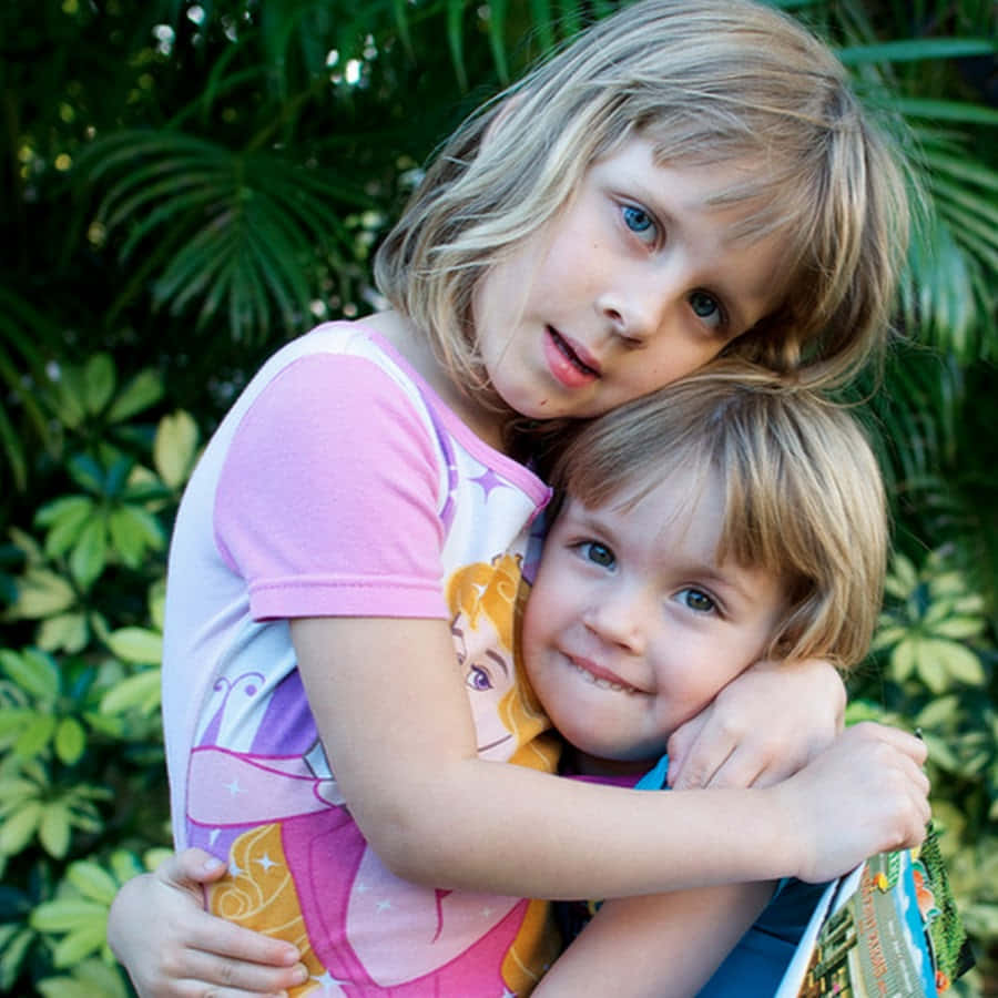 Sister 桌布 壁紙 — 兩個年幼的孩子在陽光明媚的花園裡熱情擁抱，身後是茂密的綠色樹葉。