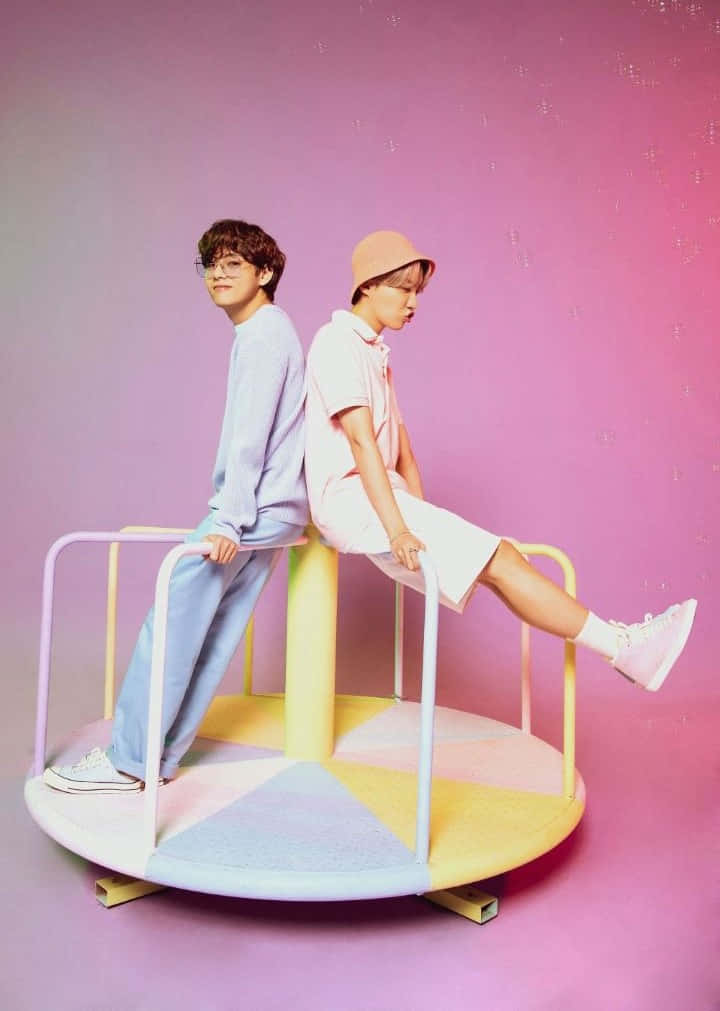 Big Hit Entertainment 桌布 壁紙 — 兩個年輕人背對背坐在色彩繽紛的旋轉木馬座位上，背景是柔和的粉紅色漸層。