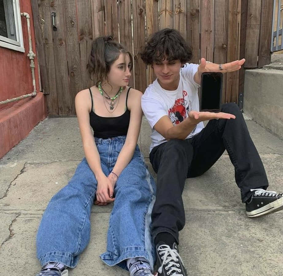 Aesthetic Couple 桌布 壁紙 — 兩個年輕人隨意坐在混凝土台階上，背後是風化的木柵欄，其中一人拿著手機當作道具。