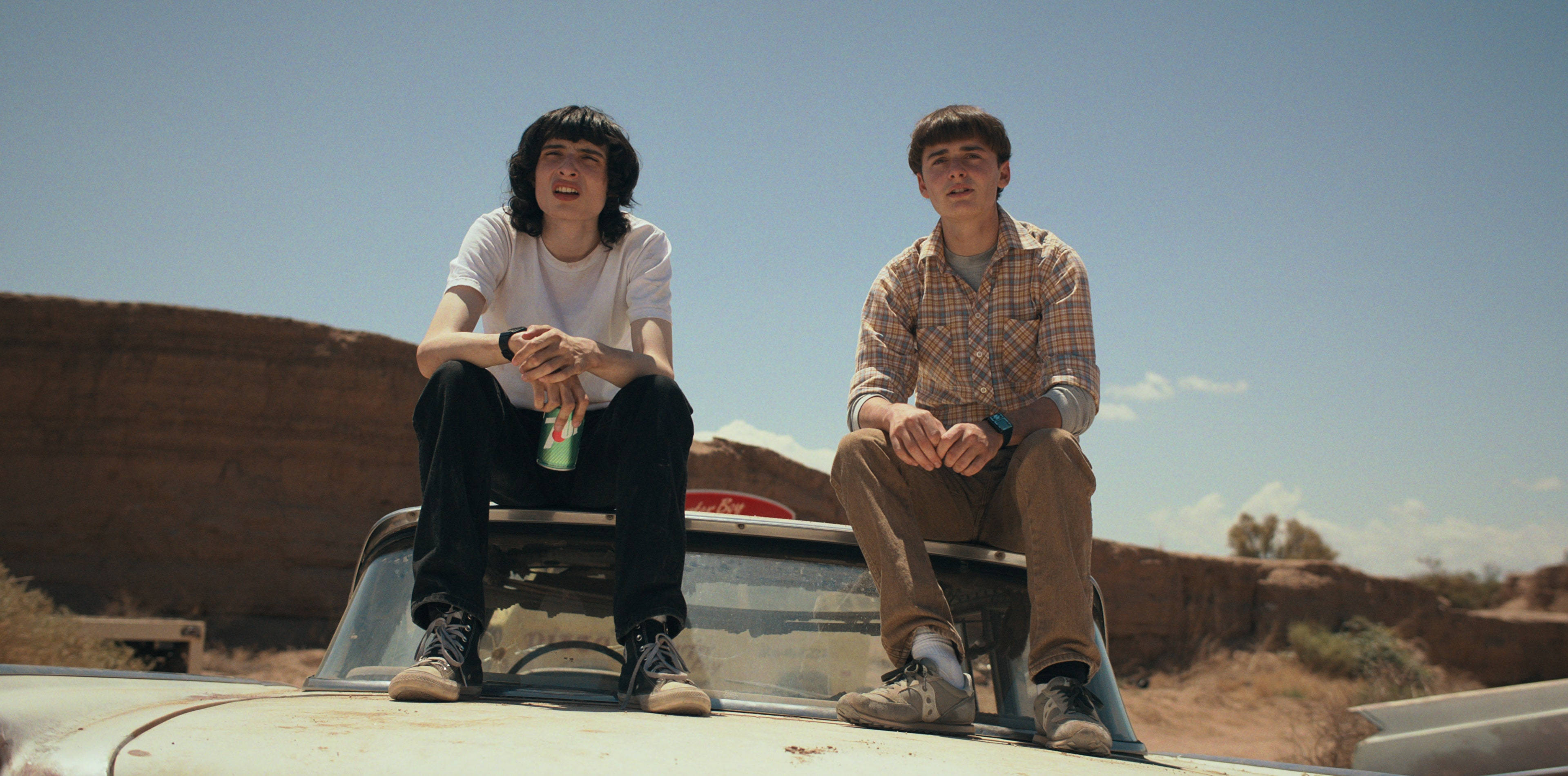 finn wolfhard 桌布 壁紙 — 兩個年輕男子坐在一輛復古車頂上，身處陽光炙烤的沙漠峽谷，頭頂是清澈的藍天。