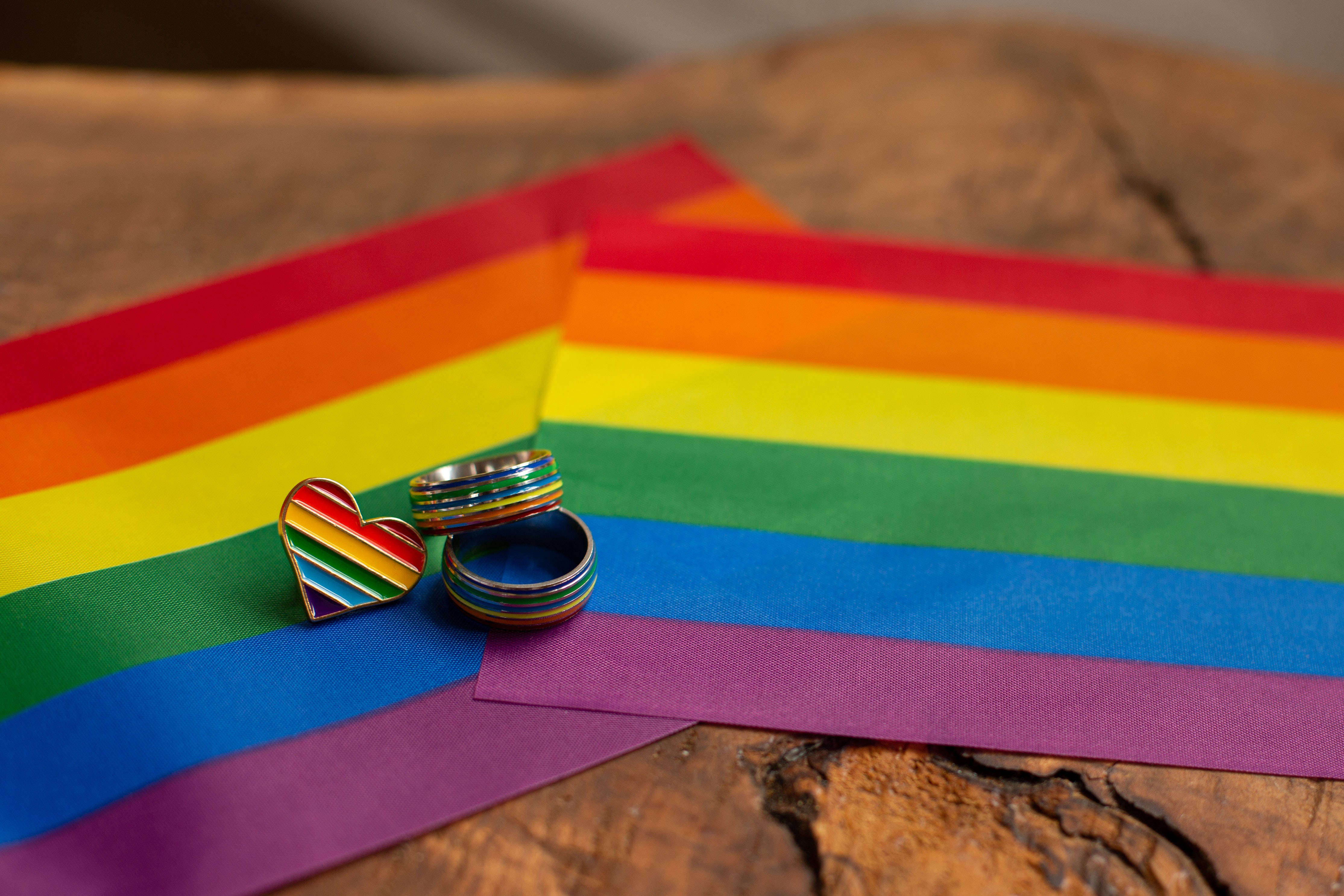 Pride Flag 桌布 壁紙 — 兩個彩虹戒指和一個心形別針放在充滿活力的彩虹旗布料上，置於質樸的木質表面。