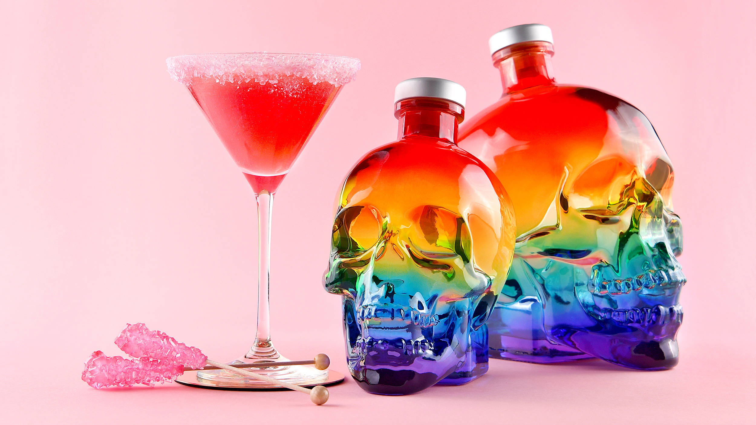 Drink 桌布 壁紙 — 兩個彩虹色骷髏形狀的酒瓶和一個糖邊玻璃杯中的紅色雞尾酒，放在柔和的粉紅色背景上。