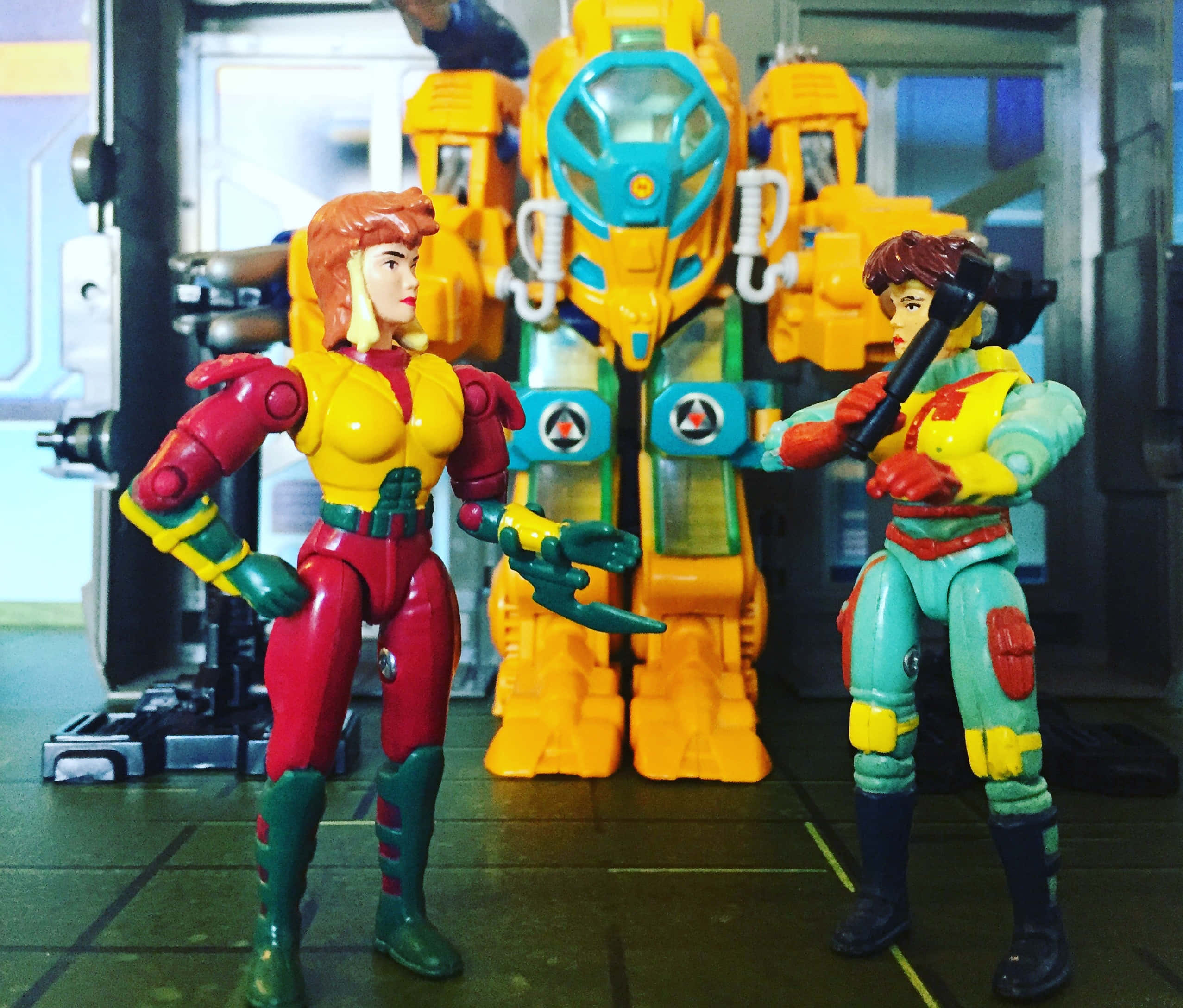 toys 桌布 壁紙 — 兩個復古動作人偶身穿色彩繽紛的服裝，在科幻機庫中手持武器，準備好面對一個巨大的黃色機器人。