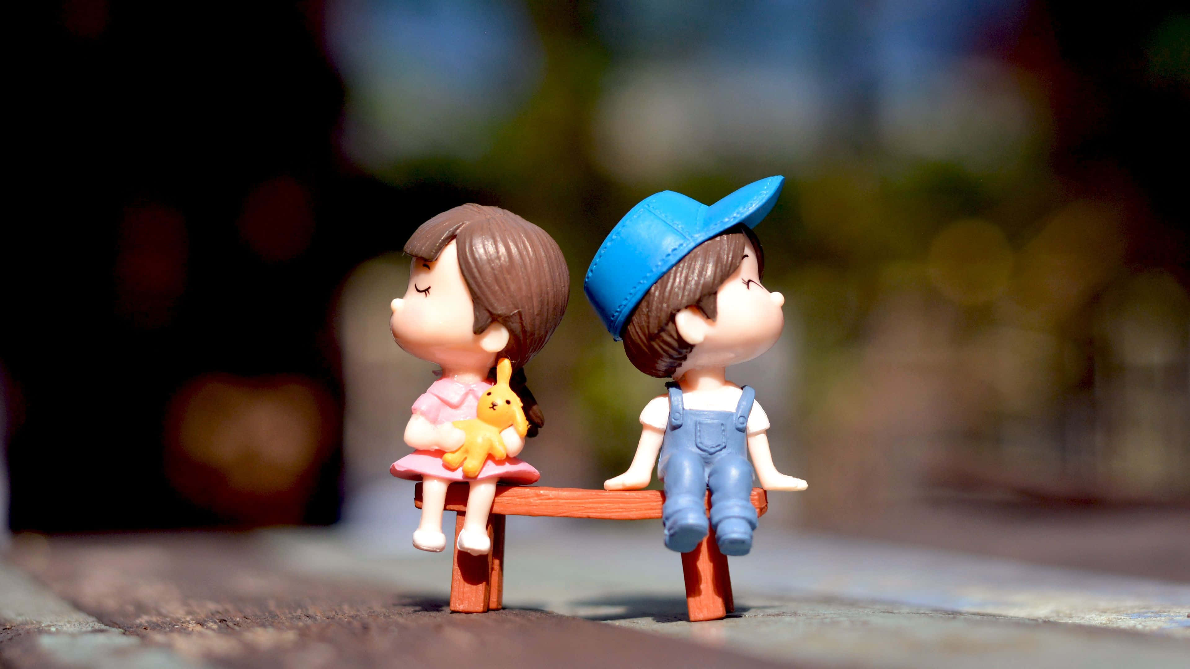 Sweet Love 桌布 壁紙 — 兩個微型玩具人物並排坐在一張小木凳上，其中一個抱著毛絨玩具，背景是柔和模糊的戶外景色。