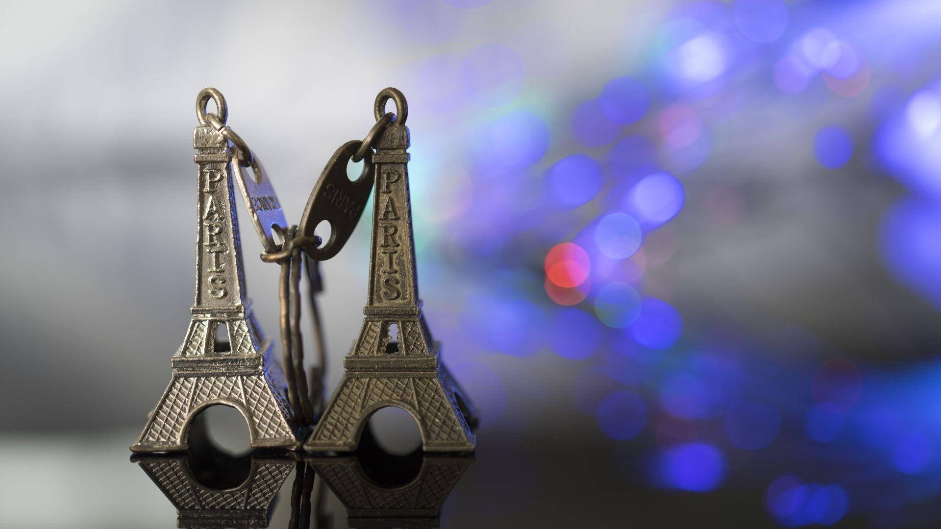 Eiffel Tower 桌布 壁紙 — 兩個微型艾菲爾鐵塔鑰匙圈掛在充滿活力的藍色散景背景上，散落著五彩繽紛的燈光。