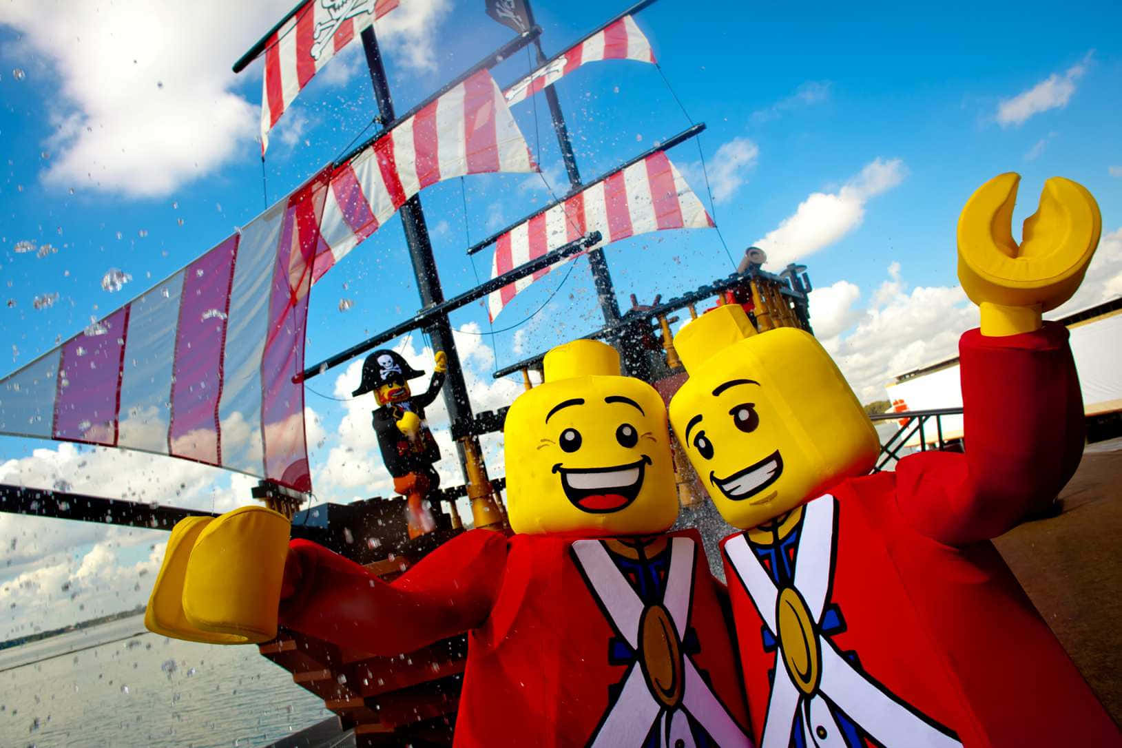 Legoland 桌布 壁紙 — 兩個微笑的LEGO海盜人偶身穿紅色外套，在藍天白雲下從一艘帶有條紋帆的船上愉快地揮手。