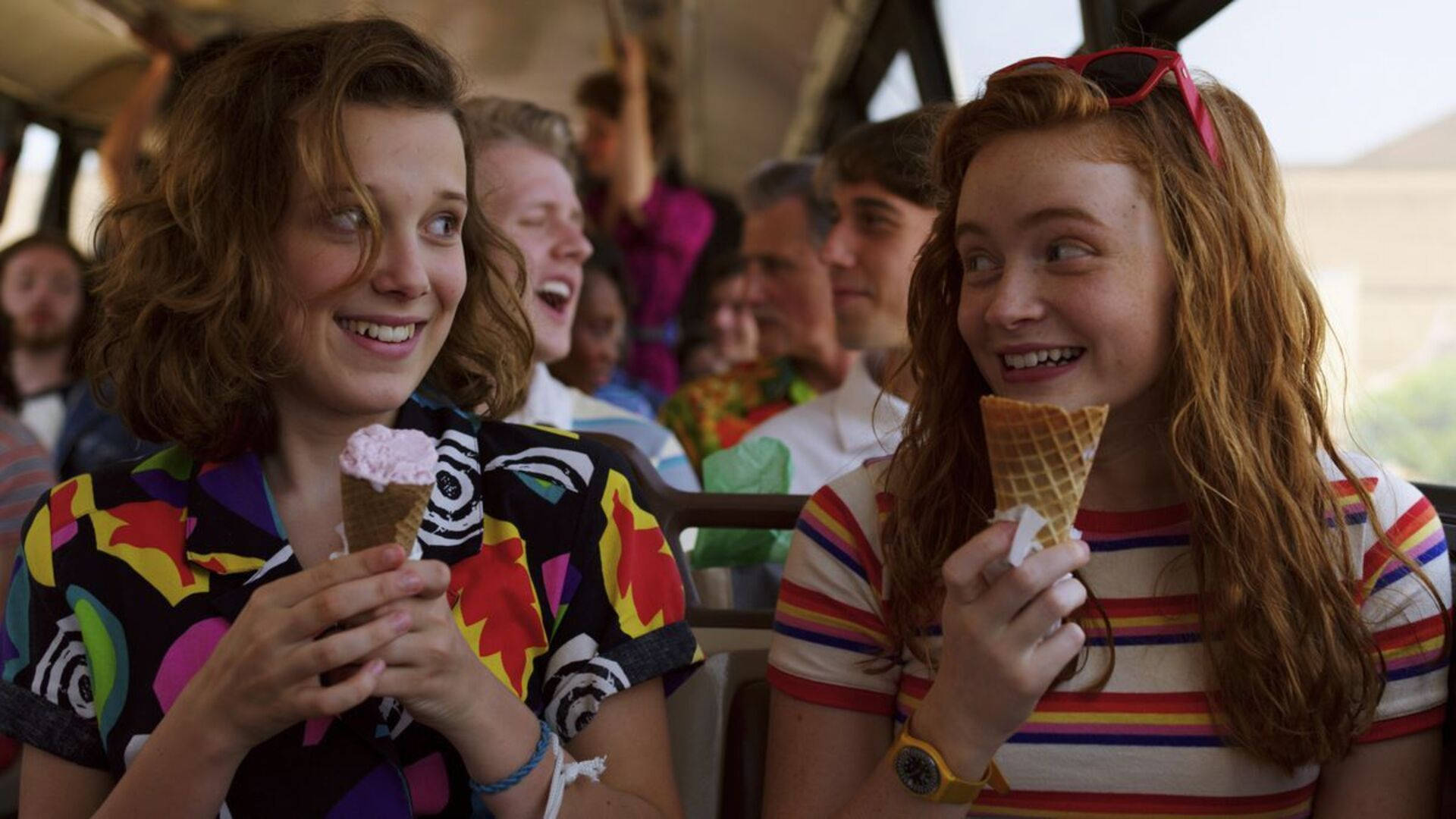 Stranger Things 3 桌布 壁紙 — 兩個微笑的女孩在陽光明媚的公車旅程中享用冰淇淋甜筒，身後有歡樂的乘客。