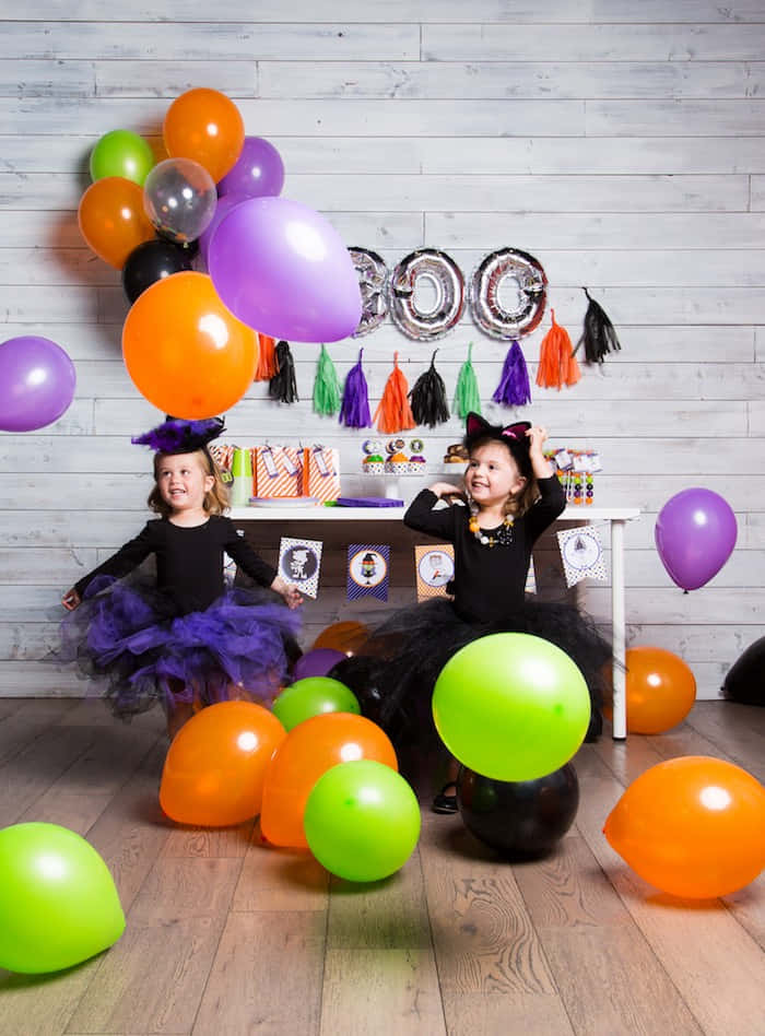 Party 桌布 壁紙 — 兩個微笑的女孩穿著女巫服裝，在色彩繽紛的萬聖節氣球和裝飾中，以木牆為背景擺姿勢。