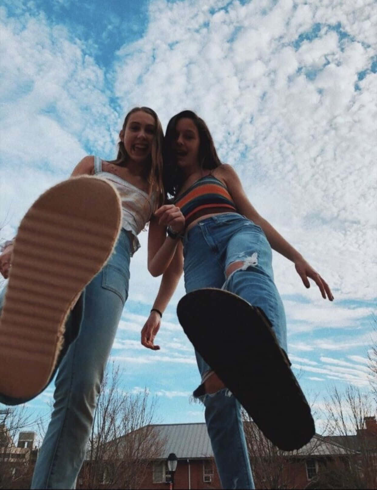 摯友 桌布 壁紙 — 兩個微笑的女孩穿著破洞牛仔褲和背心，在藍天白雲下俏皮地舉起運動鞋擺姿勢。
