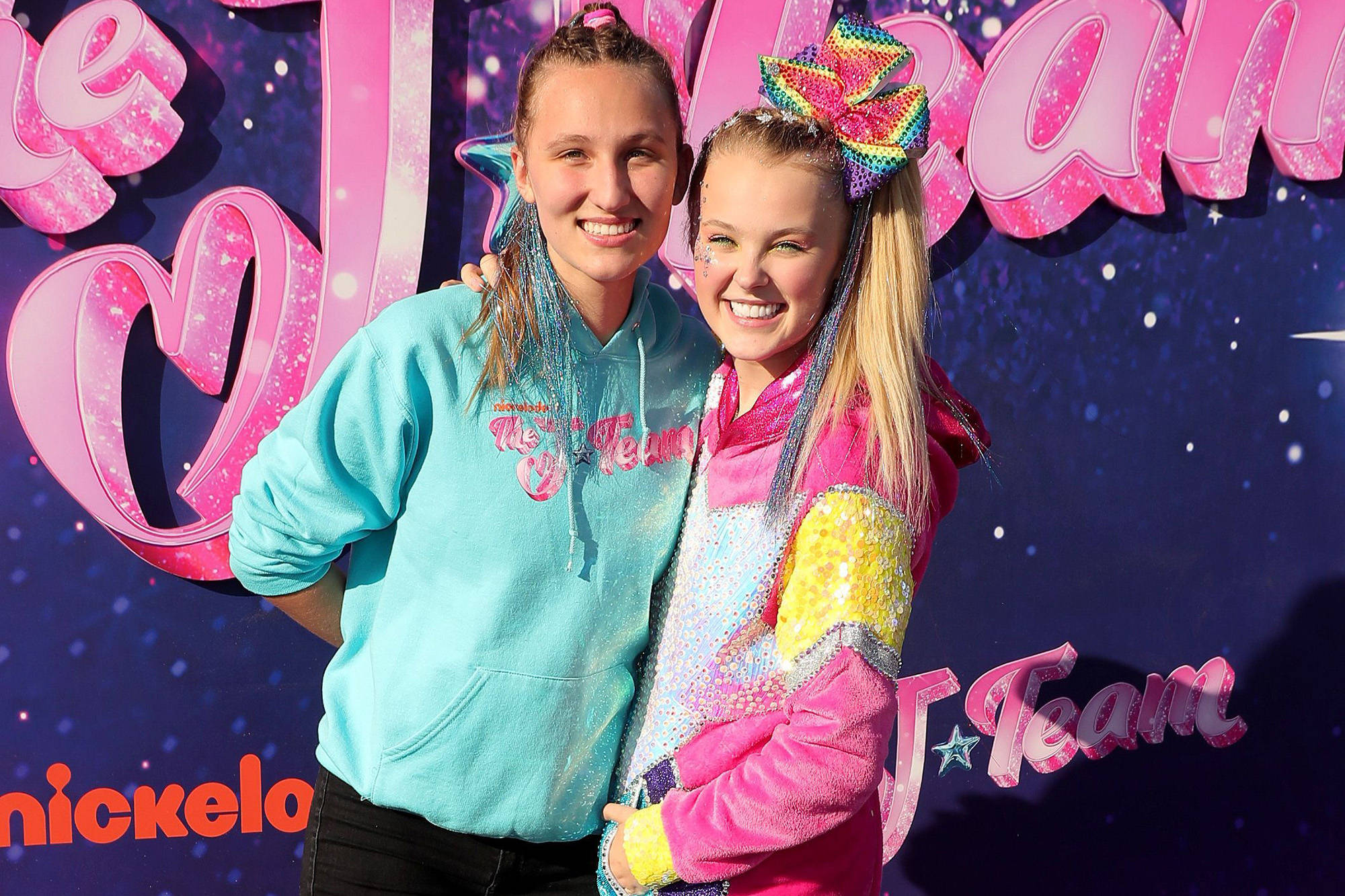 JoJo Siwa 桌布 壁紙 — 兩個微笑的女孩穿著色彩繽紛的閃亮服裝，在帶有星星的充滿活力的粉紅色和紫色 'JoJo Siwa' 品牌背景前擺姿勢。