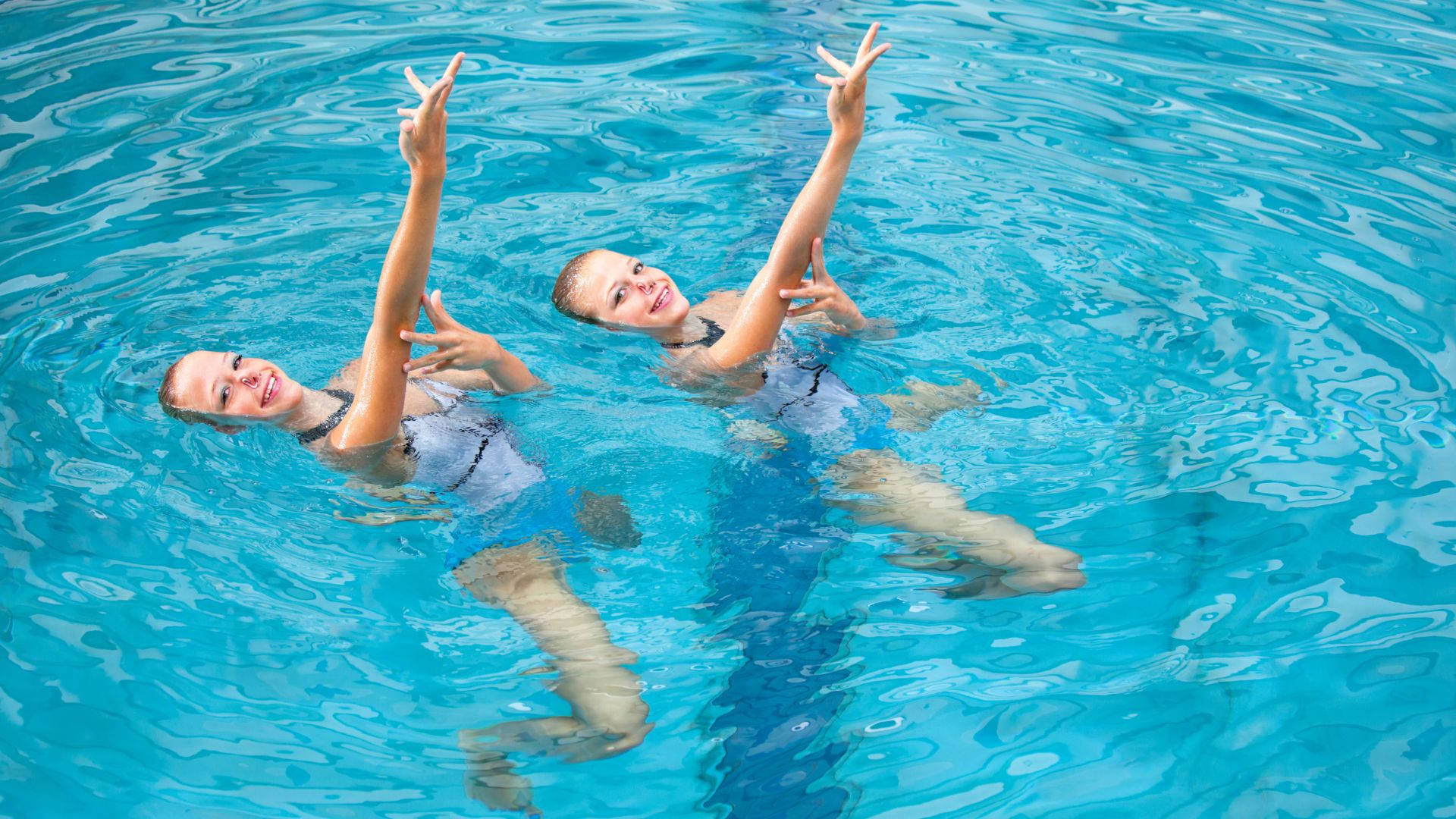 Swimming 桌布 壁紙 — 兩個微笑的女孩背浮在清澈的藍色泳池水中，舉起雙手做出和平手勢。