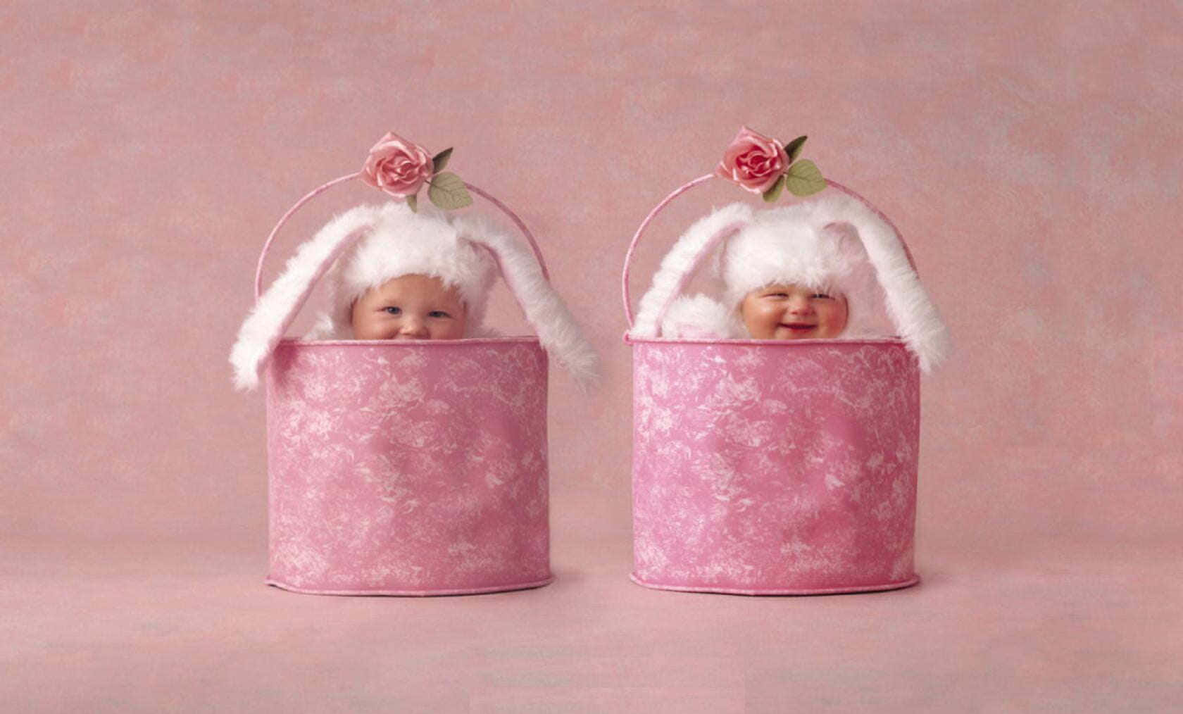 Cute Babies 桌布 壁紙 — 兩個微笑的嬰兒戴著粉紅色兔子帽，從相配的花卉圖案桶中探出頭來，背景是柔和的粉紅色。