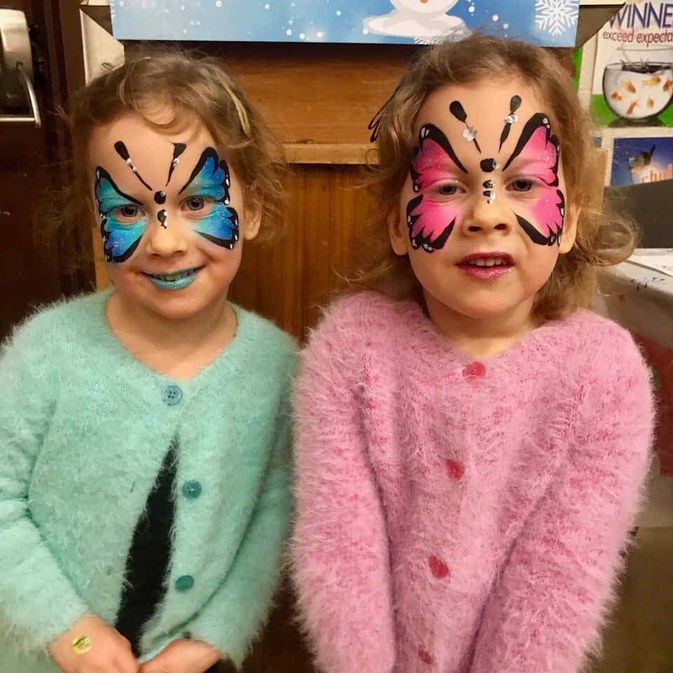 Butterfly Face Painting 桌布 壁紙 — 兩個微笑的孩子，臉上畫著蝴蝶圖案，一個藍色一個粉紅色，穿著毛絨絨的毛衣在室內。