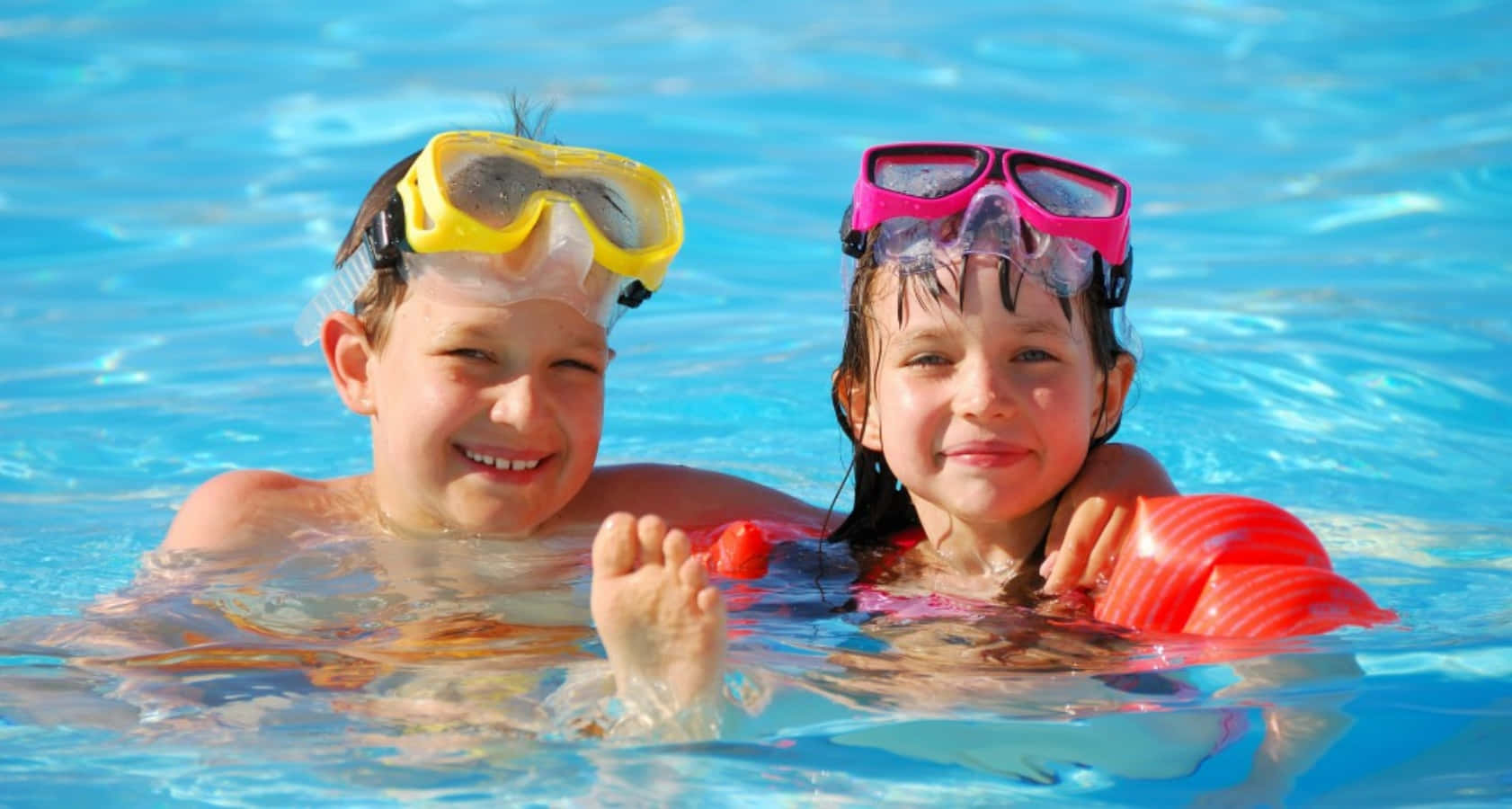 Swimming 桌布 壁紙 — 兩個微笑的孩子在明亮的藍色泳池中，戴著彩色泳鏡和浮臂。