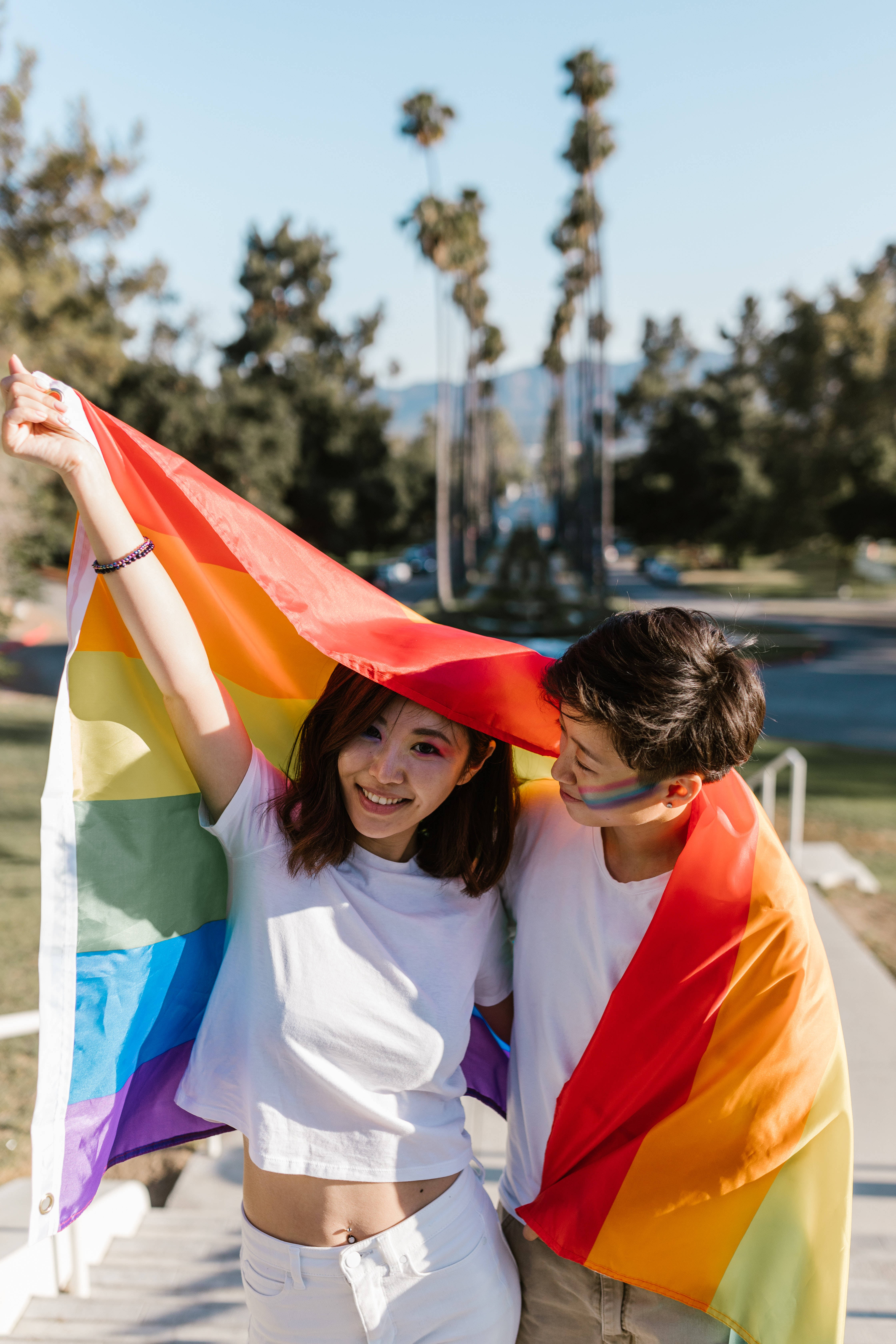 女同志 桌布 壁紙 — 兩個微笑的年輕人在陽光明媚的公園裡，在鮮豔的彩虹旗下擁抱，背景有棕櫚樹和遠山。