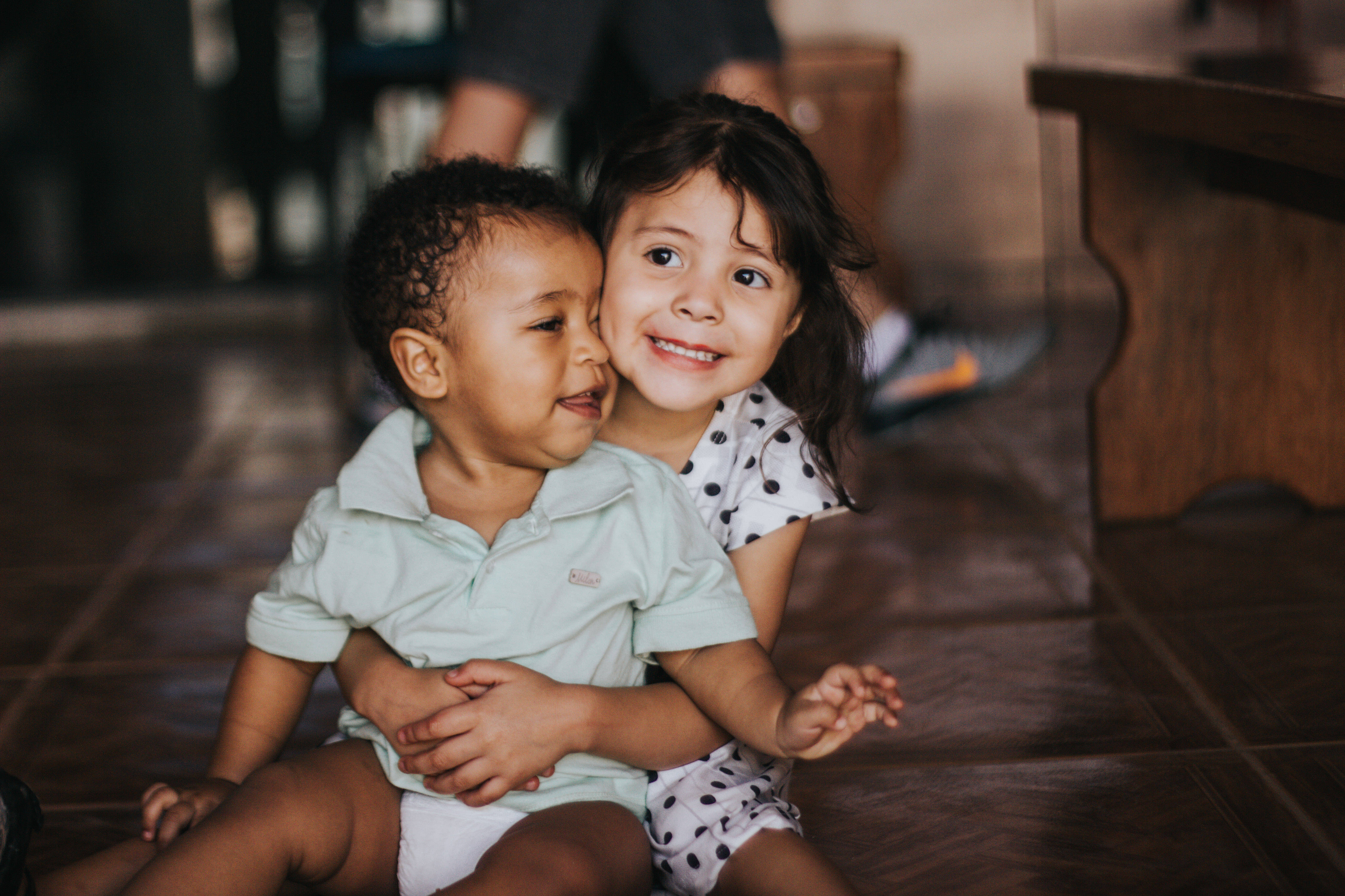 小孩動圖 桌布 壁紙 — 兩個微笑的幼兒在光線柔和的室內木地板上溫暖地擁抱。