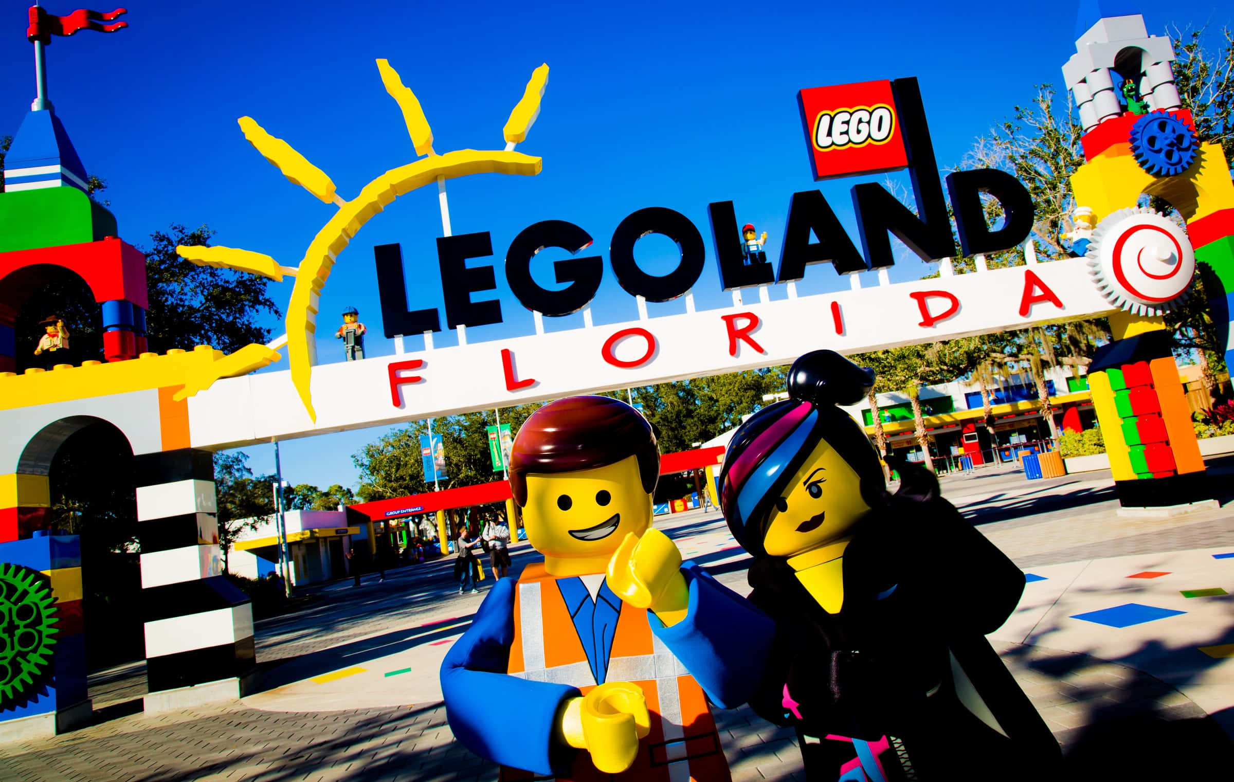 Legoland 桌布 壁紙 — 兩個微笑的樂高迷你人偶在晴朗的日子裡，在色彩繽紛的樂高樂園佛羅里達入口拱門下愉快地擺姿勢。