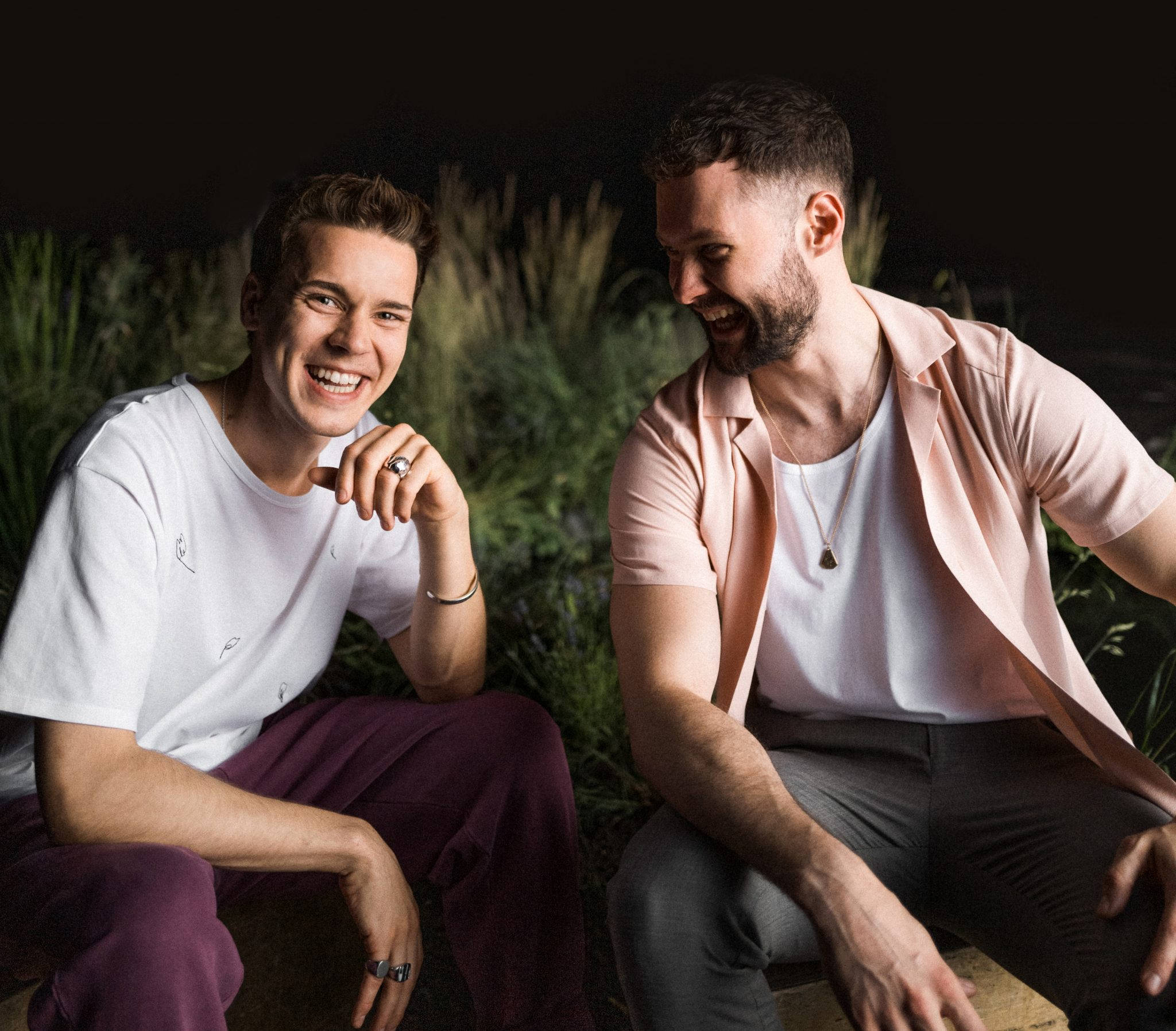 Calum Scott 桌布 壁紙 — 兩個微笑的男人在戶外夜晚被高大的綠色植物包圍，分享歡樂的時刻。