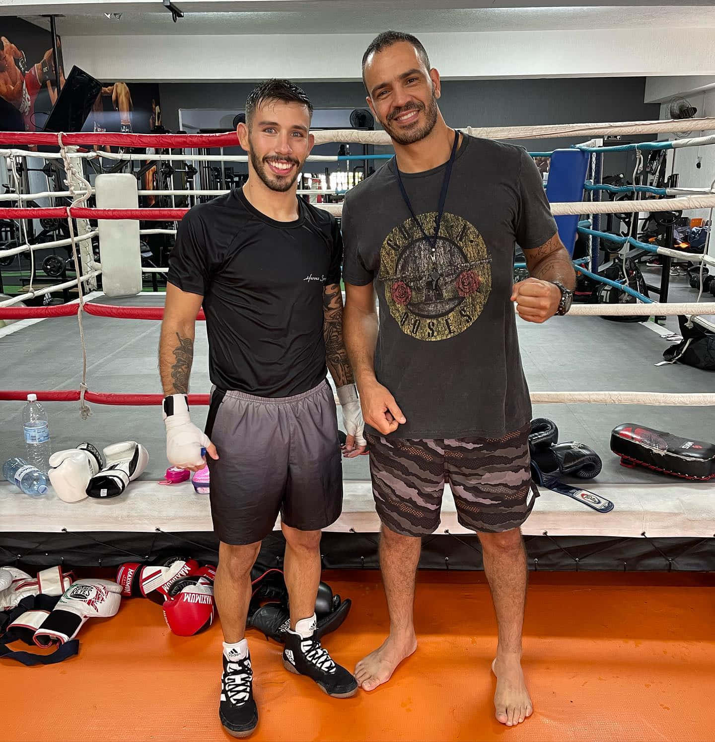 MMA 桌布 壁紙 — 兩個微笑的男人在拳擊擂台上自信地擺姿勢，周圍是訓練裝備和健身器材。