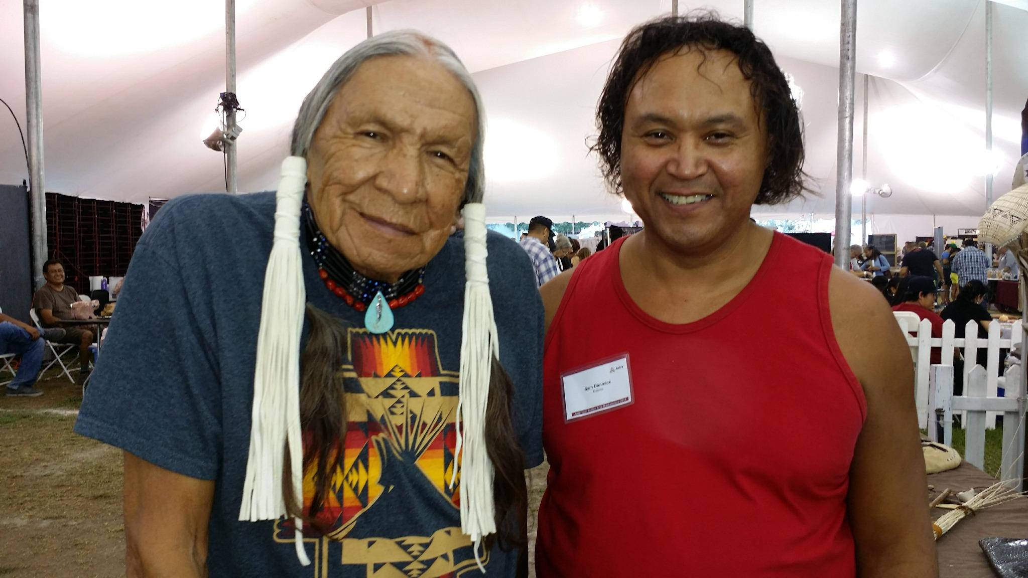 Native American 桌布 壁紙 — 兩個微笑的男人在白色帳篷下的戶外活動中一起擺姿勢。一個穿著帶有流蘇和珠飾的傳統美洲原住民服裝，另一個穿著紅色背心。