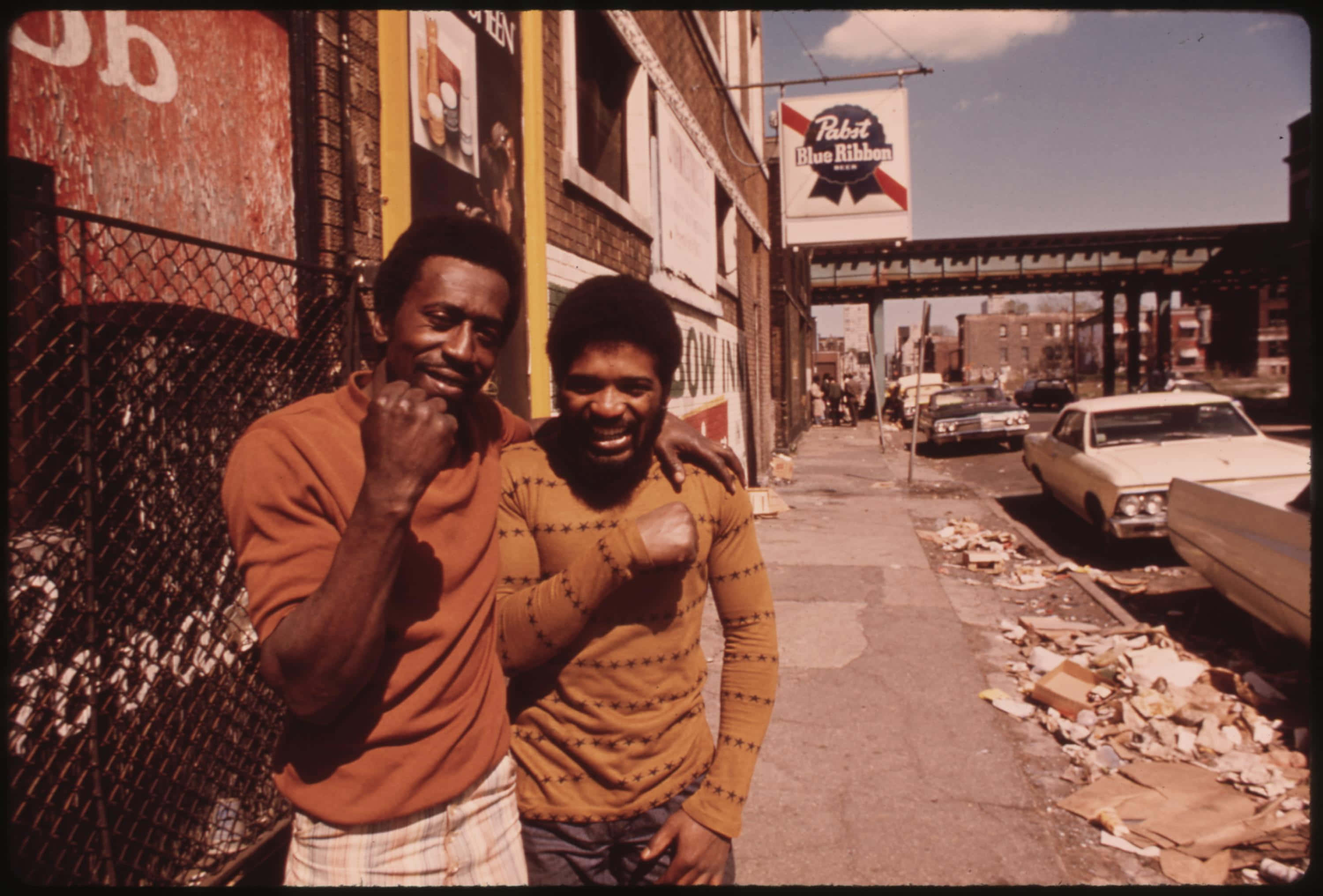 1970s 桌布 壁紙 — 兩個微笑的男人在陽光明媚的城市街道上頑皮地擺姿勢，頭頂上有復古汽車和Pabst Blue Ribbon招牌。