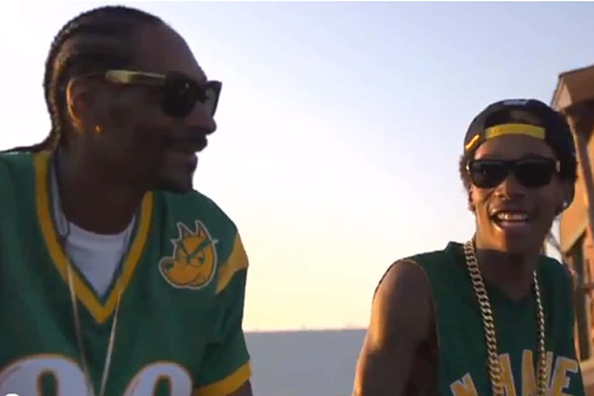 Young Snoop Dogg 桌布 壁紙 — 兩個微笑的男人穿著綠色球衣和太陽眼鏡，在朦朧的日落天空下一起大笑。