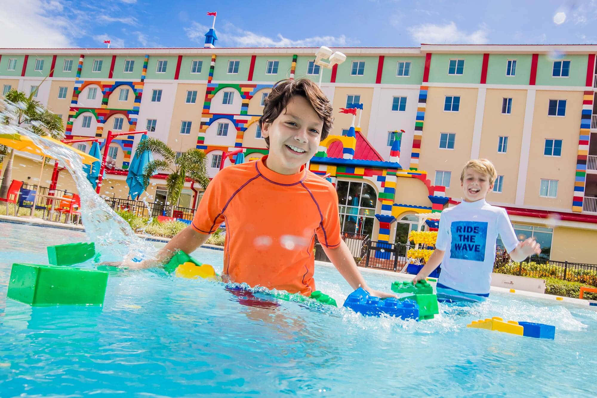 Legoland 桌布 壁紙 — 兩個微笑的男孩在明亮的藍色泳池中玩巨型樂高積木，在陽光明媚的天空下濺起水花。